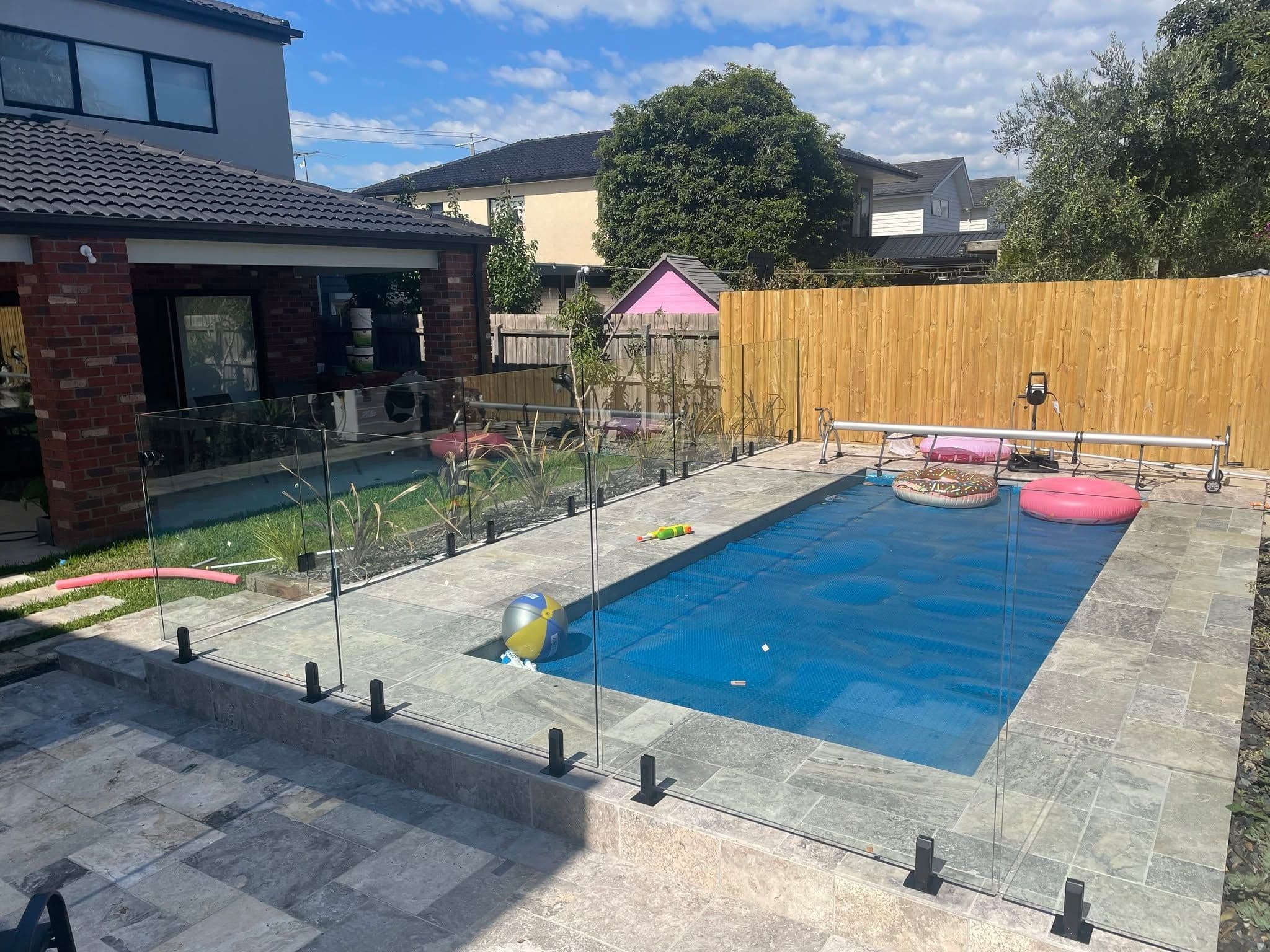Frameless glass pool fence with matte black square spigots core-drilled into stone coping surrounding a backyard swimming pool in a suburban Melbourne property.
