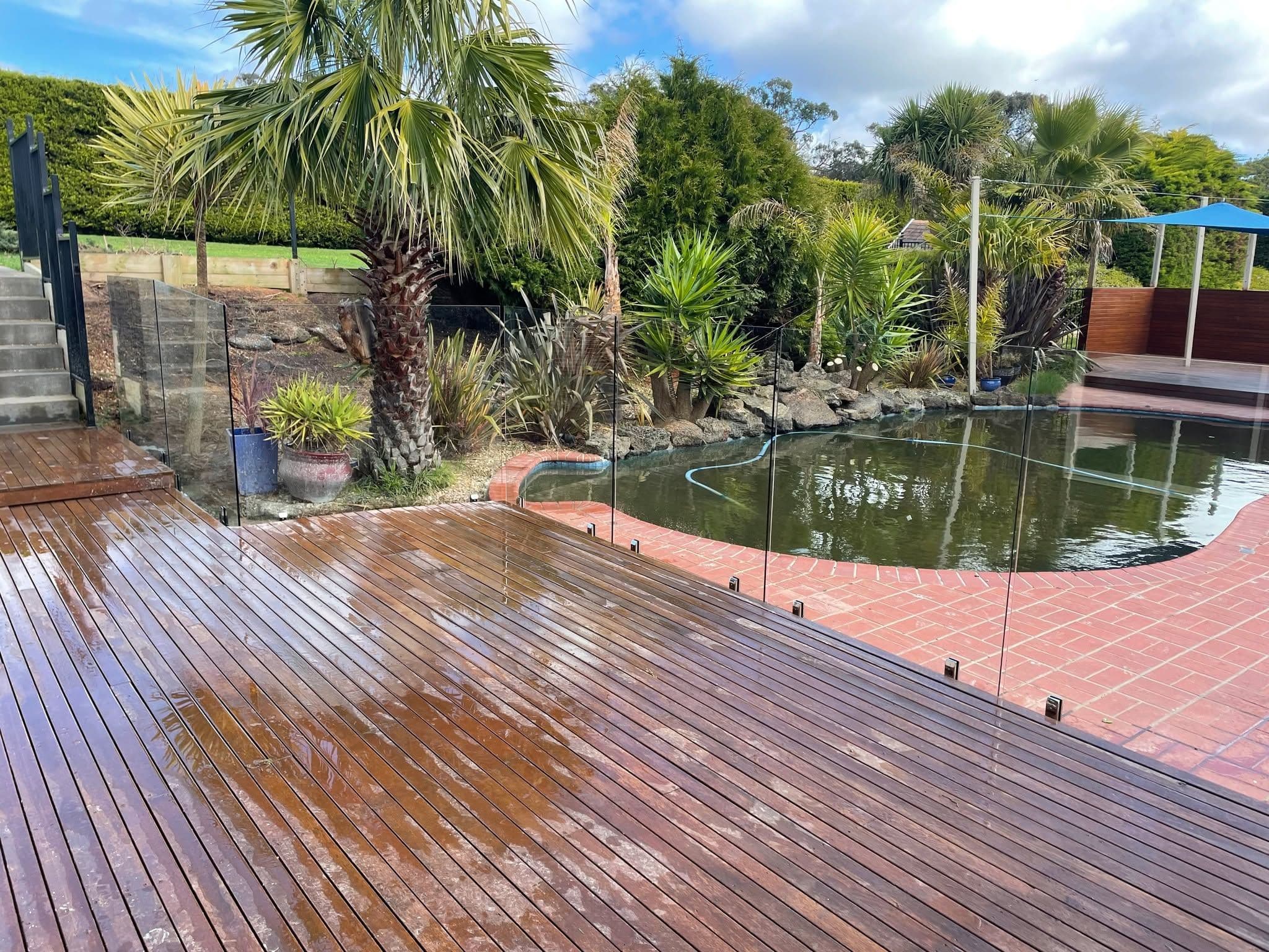 Frameless glass pool fence with matte black square spigots core-drilled into red brick coping, enclosing a freeform backyard pool beside a timber deck.