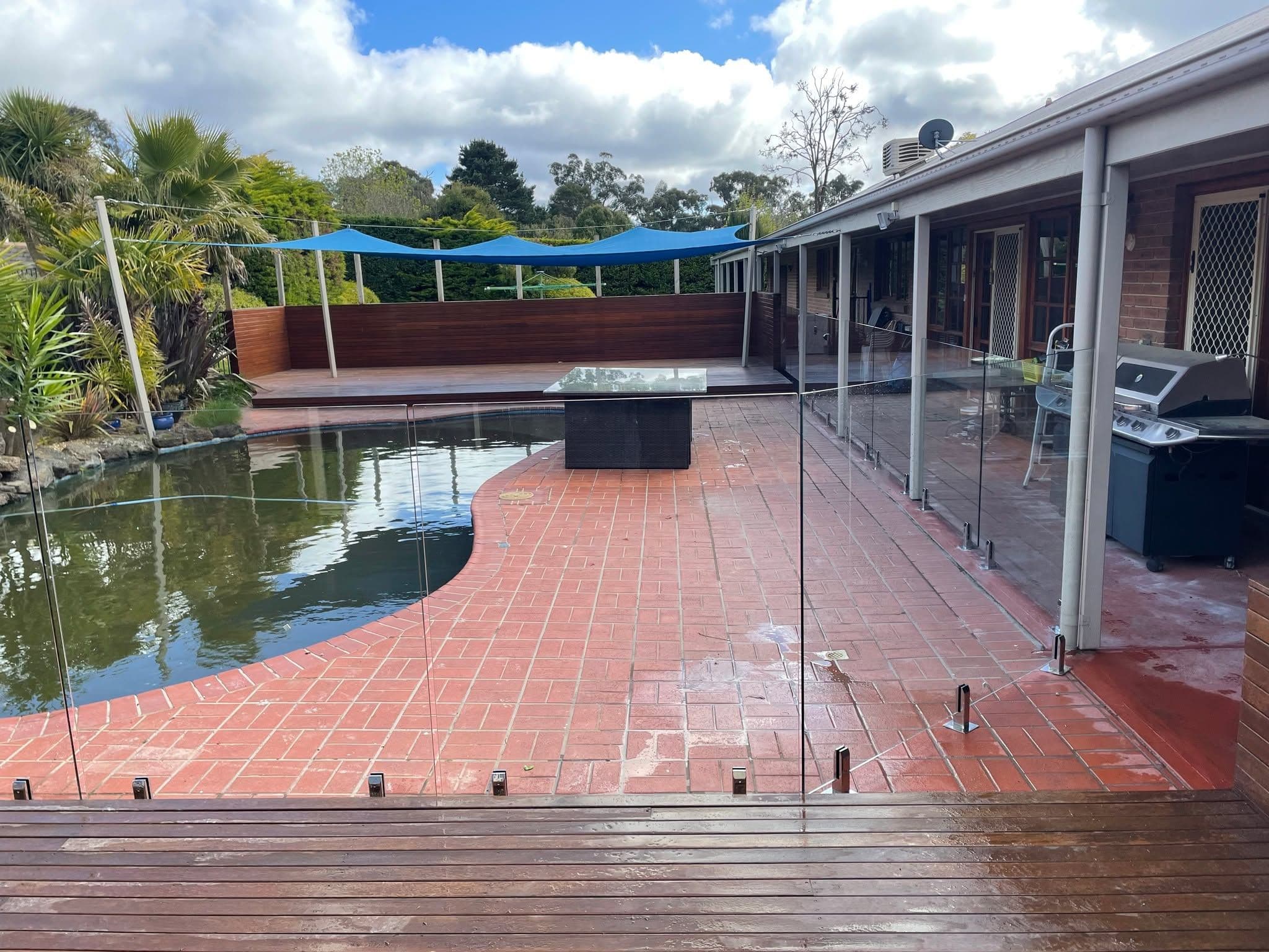 Frameless glass pool fence with chrome spigots core-drilled into brick paving and timber decking, surrounding a freeform backyard swimming pool with a covered alfresco entertaining area and timber deck beyond.