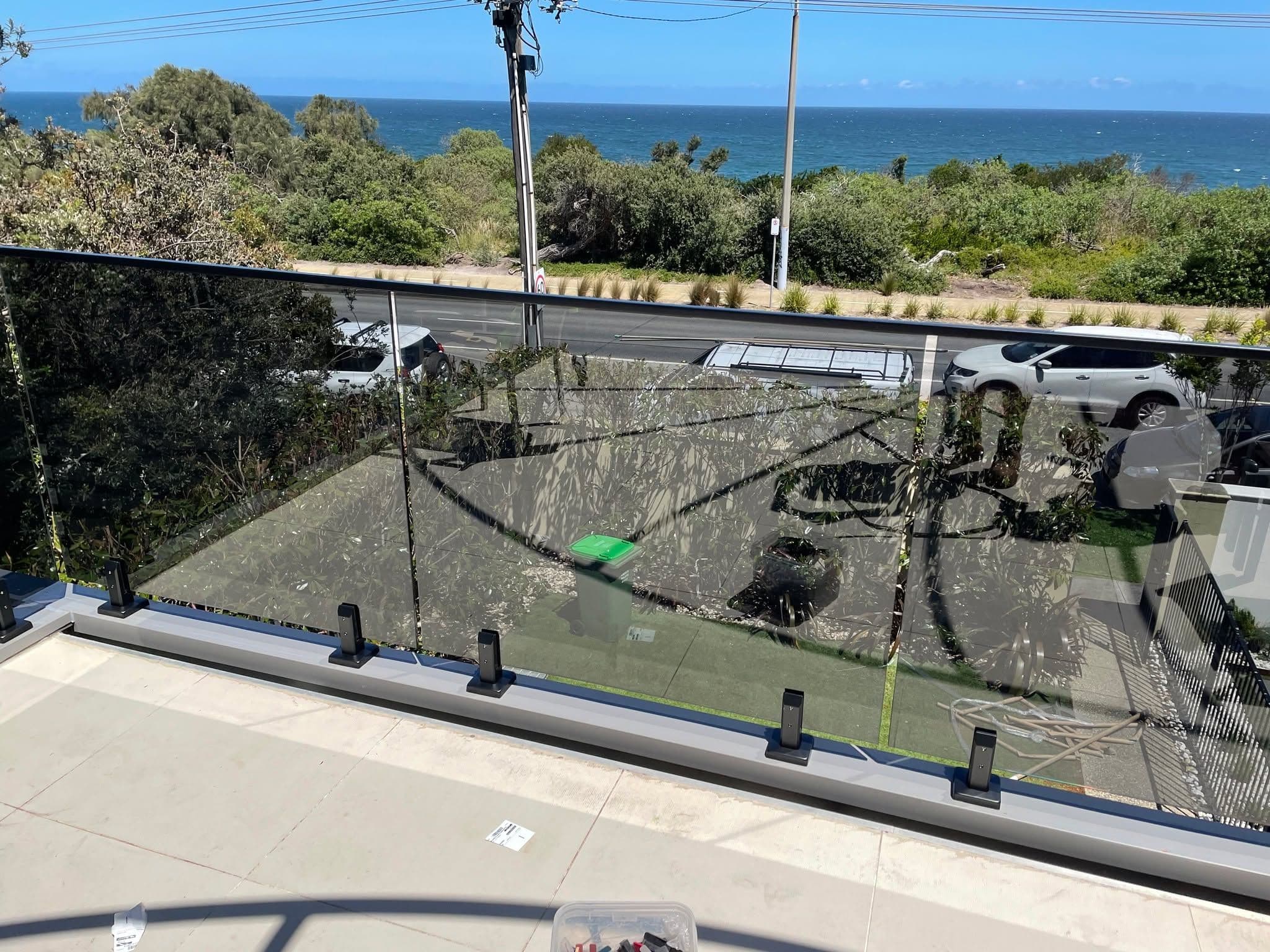 Clear glass balustrade with matte black spigots and a top aluminium handrail channel on an elevated coastal balcony overlooking the ocean.