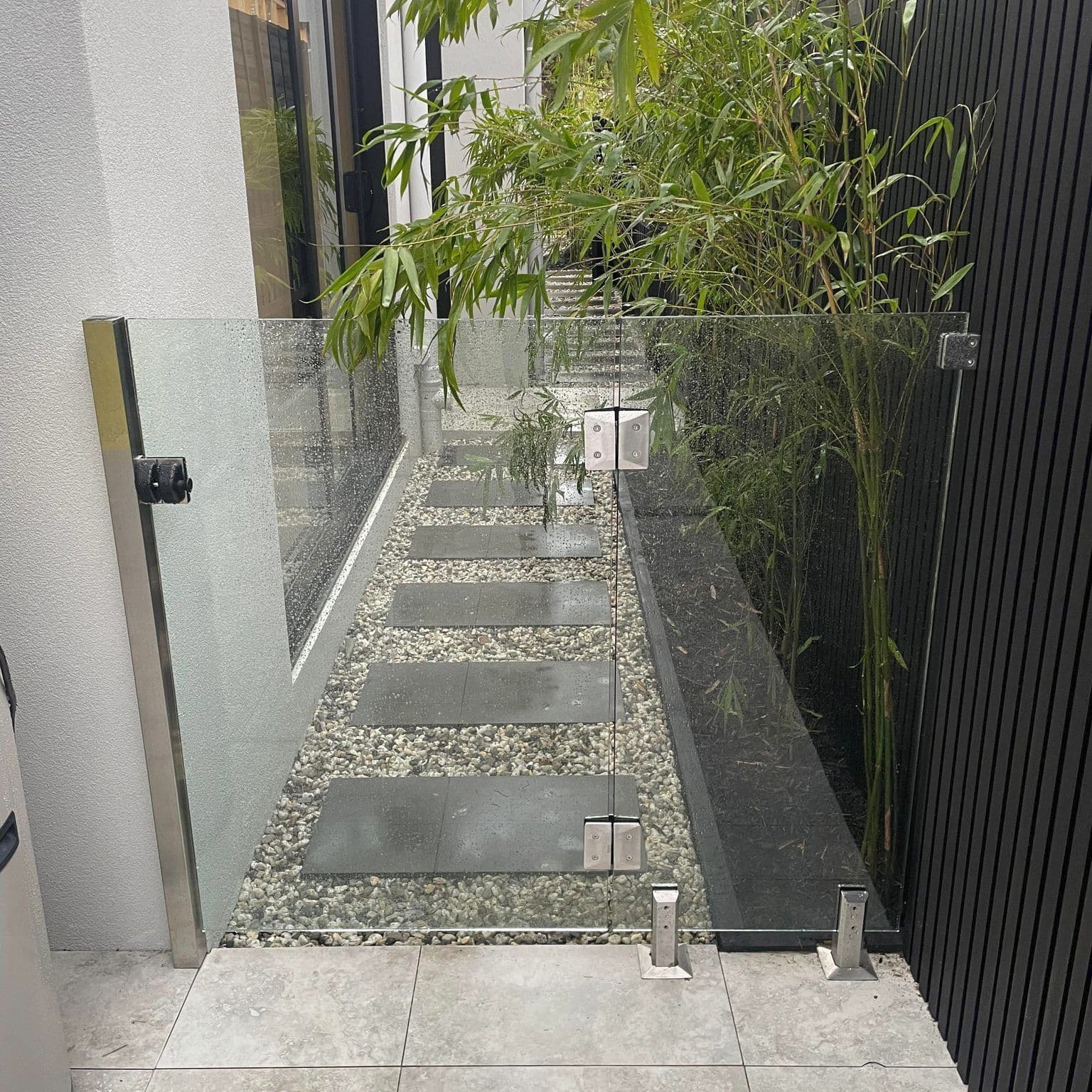 Frameless glass gate or balustrade panel with brushed nickel square spigots and wall-mounted hardware, installed at the entry of an outdoor garden pathway with stepping stones, pebbles, and bamboo plants.