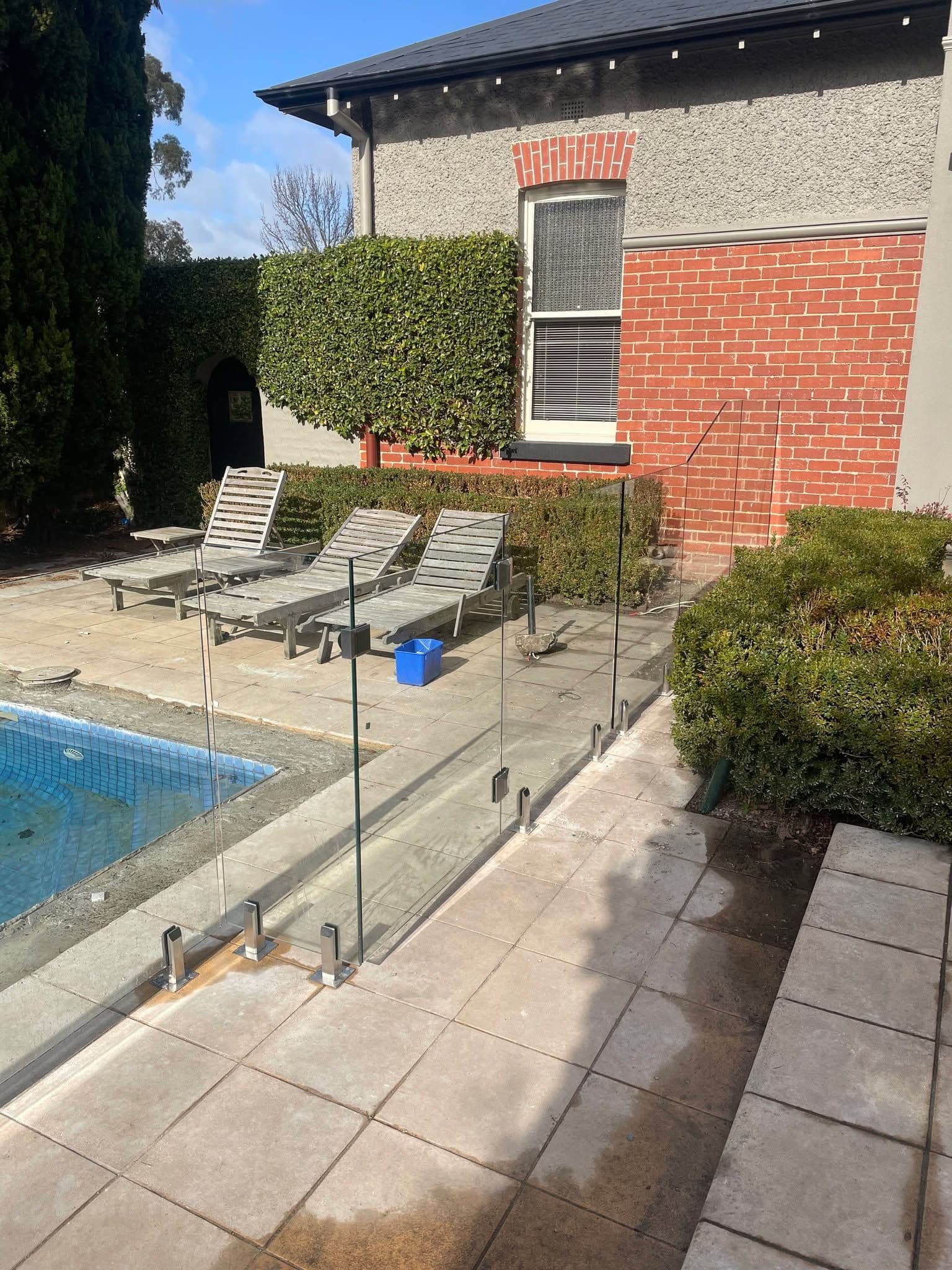 Frameless glass pool fence with square chrome spigots core-drilled into tiled coping surrounding a backyard swimming pool beside a classic brick home.