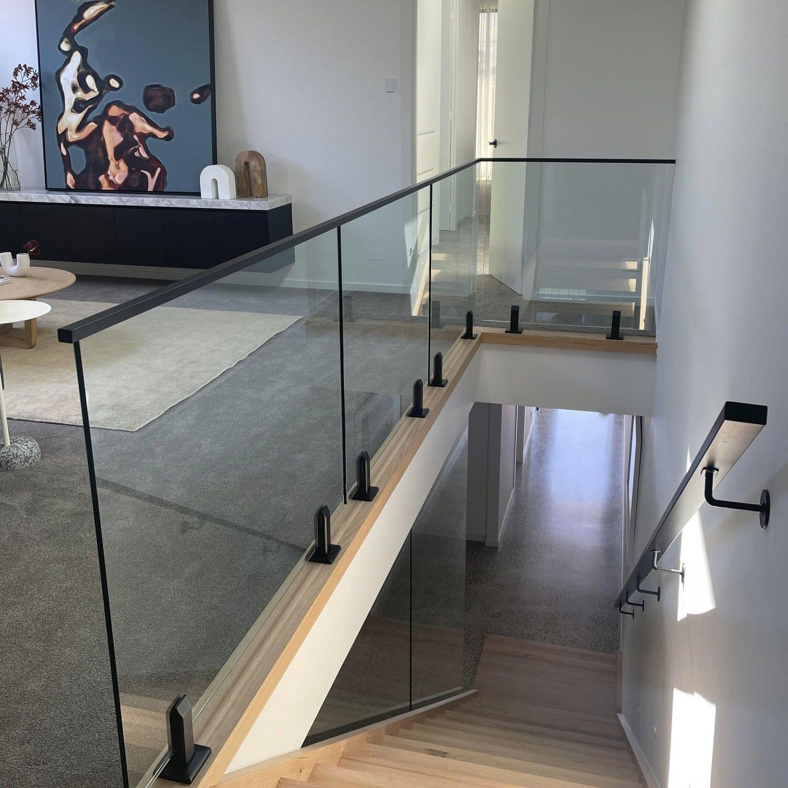 Clear glass balustrade with matte black spigots and matte black handrail lining a timber staircase and upper mezzanine landing in a contemporary open-plan home interior.