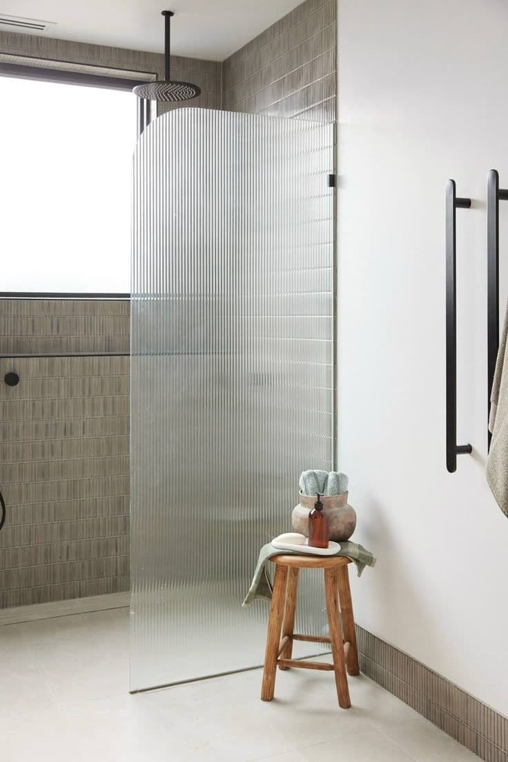Frameless fixed shower panel in reeded/fluted low iron glass with matte black hardware in a modern tiled bathroom with ceiling-mounted rain shower head.