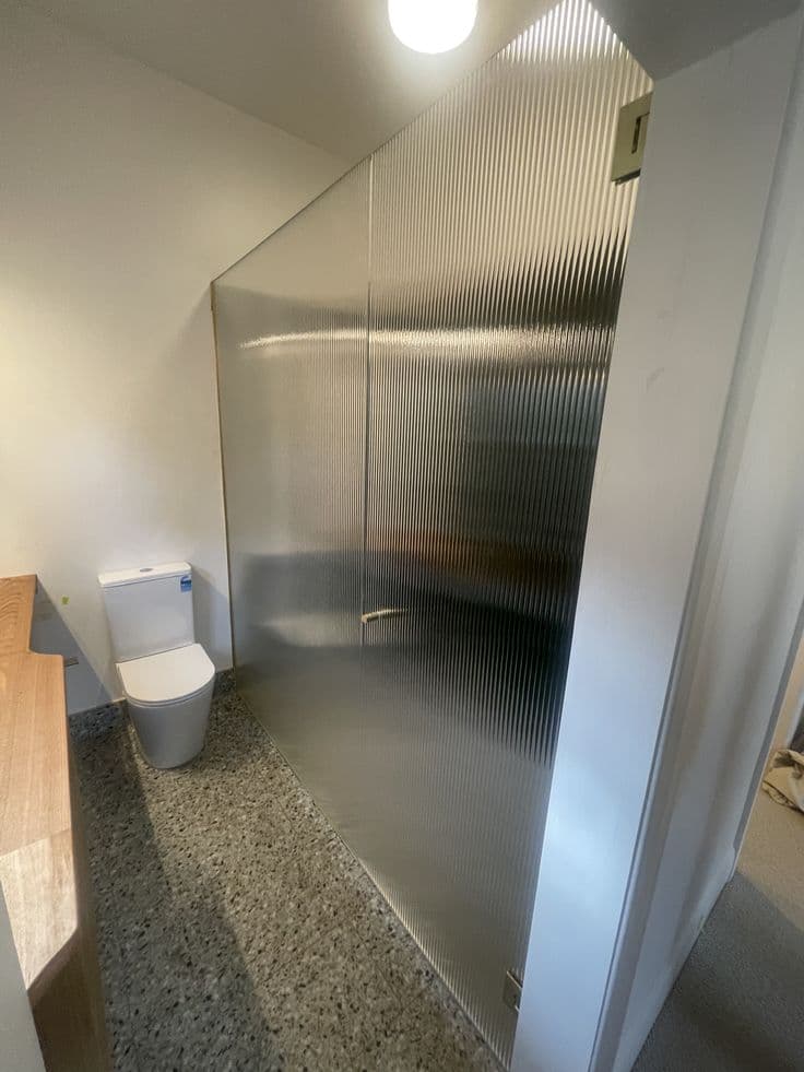 Frameless shower enclosure with reeded (narrow reed) low iron glass panel and door in a compact bathroom with terrazzo flooring and a raked ceiling, with hinges directly on the glass edge and no visible aluminium framing.