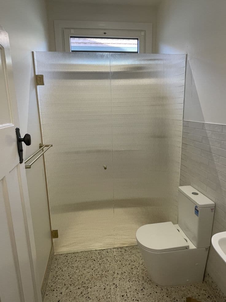 Frameless shower screen in reeded glass with brushed gold hinges and a single front-return panel configuration in a compact tiled bathroom with terrazzo flooring.