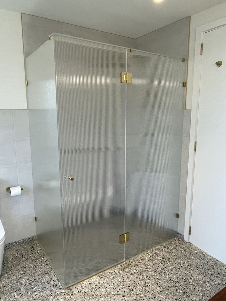 Frameless shower enclosure in a front-return configuration with reeded low iron glass panels, brushed gold square hinges and clips mounted directly on the glass, set in a light-tiled bathroom with terrazzo flooring.
