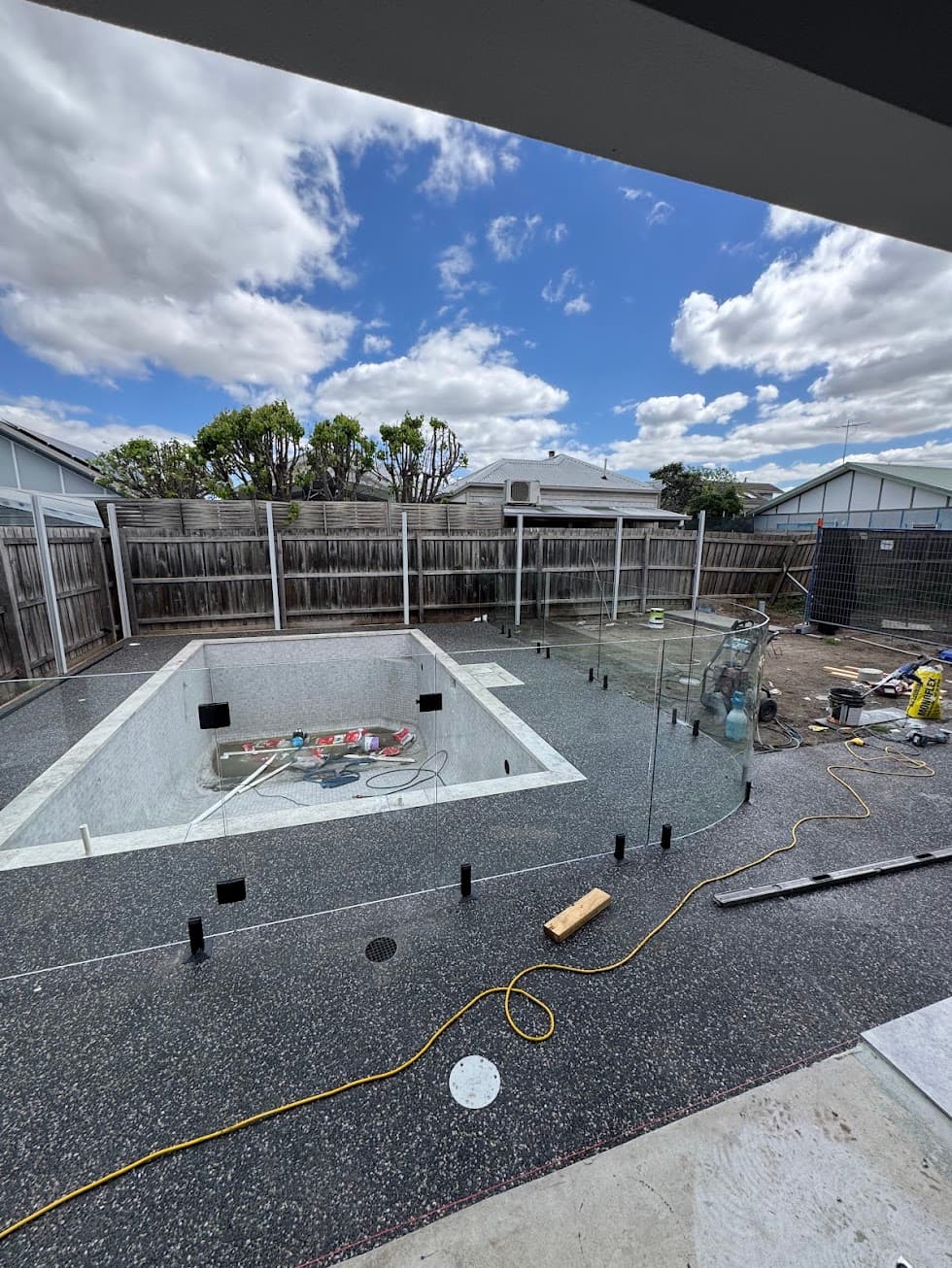Frameless glass pool fence under construction around an empty in-ground pool, with black square spigots core-drilled into aggregate coping and partially installed clear glass panels visible on site.