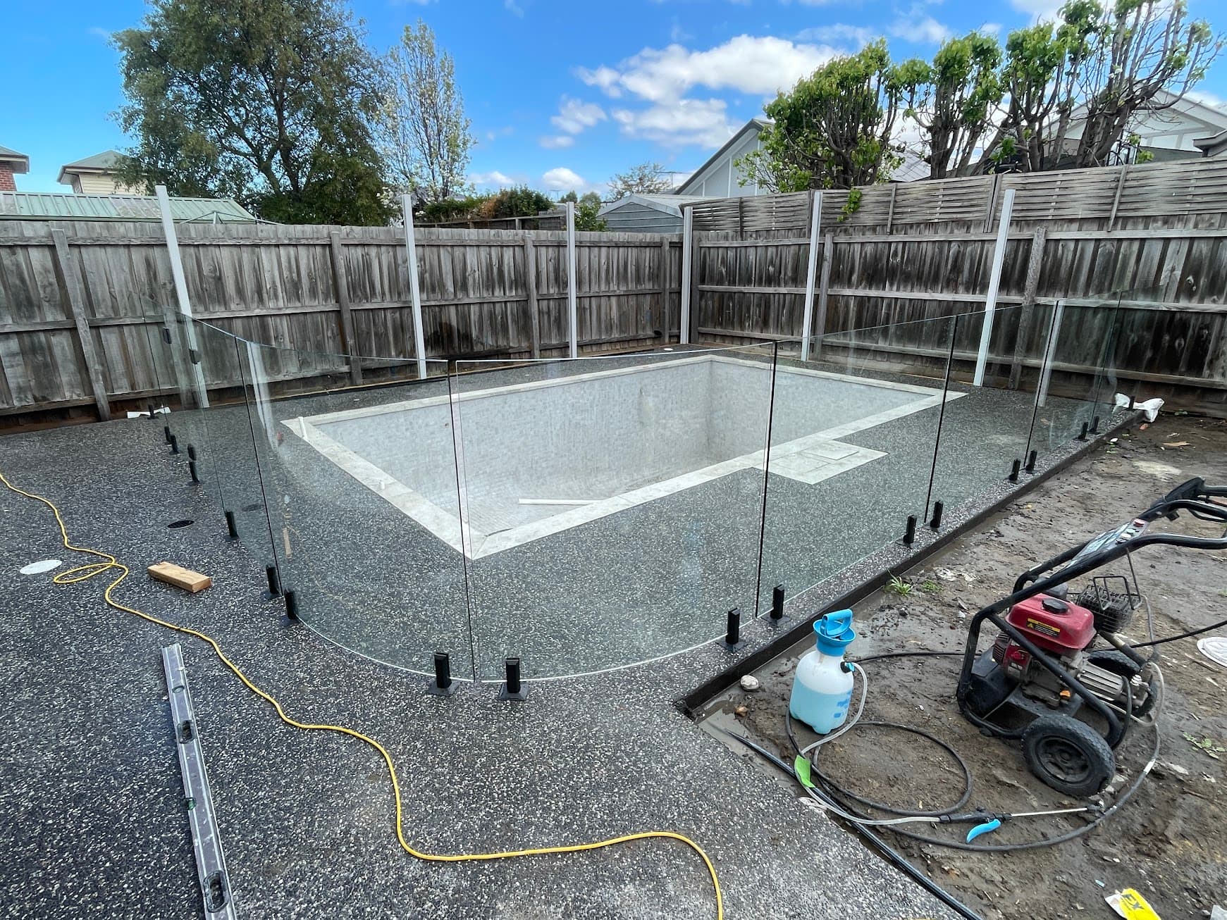 Frameless glass pool fence with matte black spigots installed around a newly constructed rectangular pool with curved corner panels, set in a suburban backyard during construction phase.