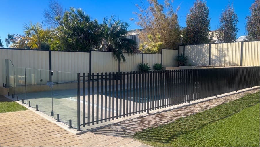 Frameless glass pool fence panels with matte black spigots installed alongside a matte black aluminium pool barrier fence surrounding a backyard pool area with brick paving.