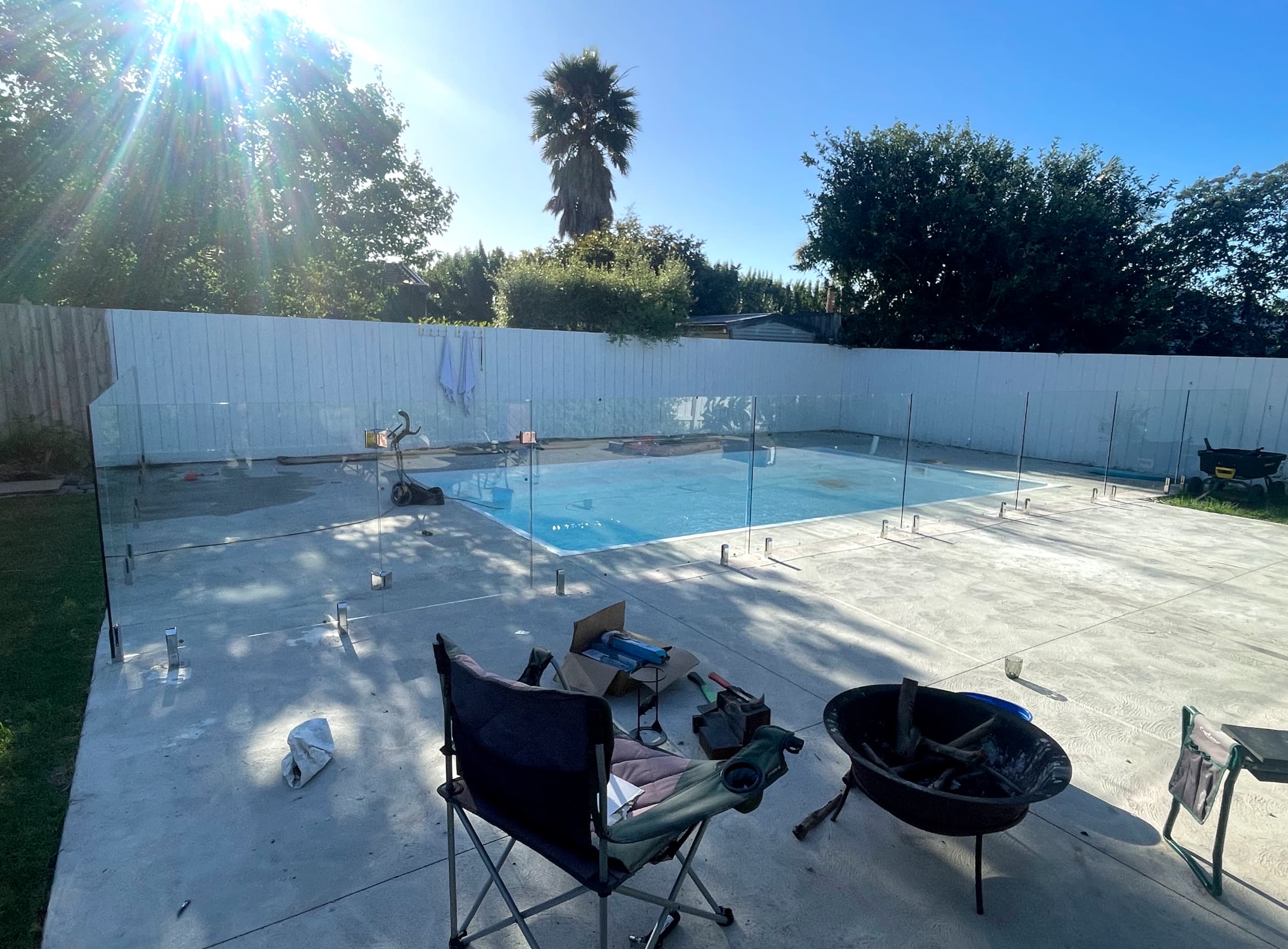 Frameless glass pool fence with chrome spigots core-drilled into concrete coping surrounding a backyard swimming pool, partially installed with panels in place and installation equipment visible on the patio.