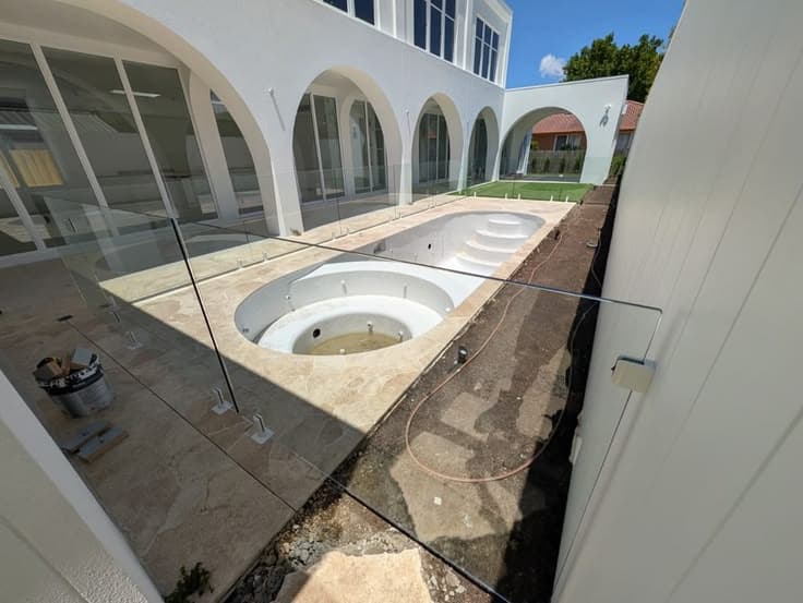 Frameless pool fence with chrome spigots core-drilled into travertine coping surrounding a newly constructed pool and spa in a luxury white rendered courtyard.
