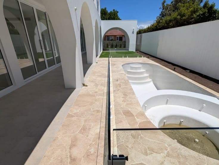 Frameless pool fence with matte black spigots core-drilled into natural stone coping surrounding a newly constructed backyard pool with entry steps.
