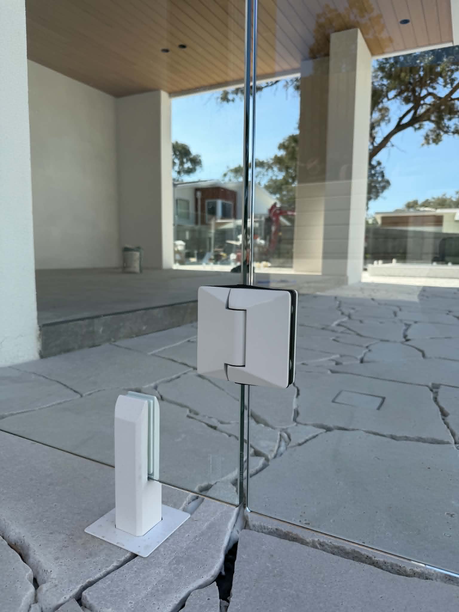 Frameless glass pool fence or balustrade with white square spigots and white glass clamp hardware on natural stone paving at a modern residential exterior.