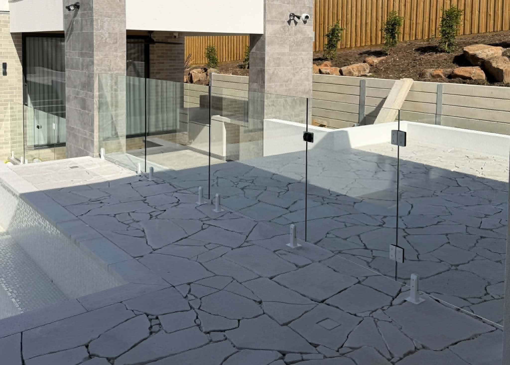 Frameless pool fence with chrome spigots core-drilled into irregular stone coping surrounding a backyard swimming pool, with clear glass panels beside a contemporary rendered home exterior.