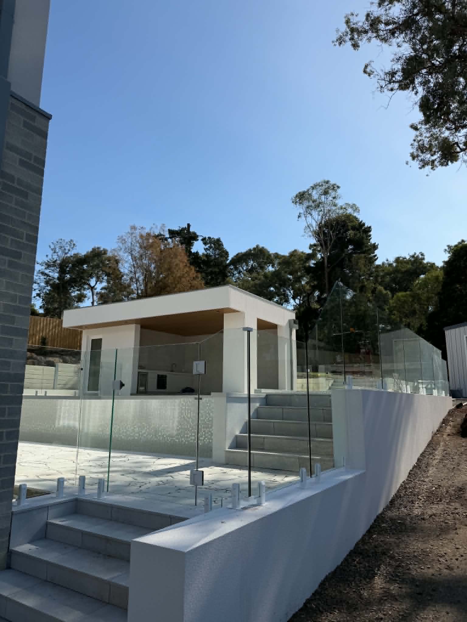 Frameless pool fence with chrome spigots core-drilled into a white rendered coping wall surrounding a residential pool area, with an alfresco cabana structure visible in the background.