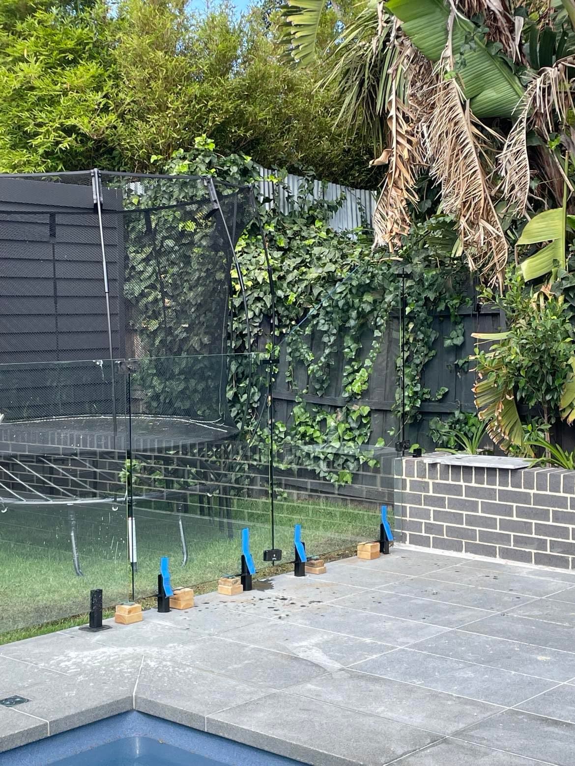 Frameless pool fence with matte black spigots core-drilled into paved coping surrounding a backyard pool, with clear glass panels still protected by blue installation tape.