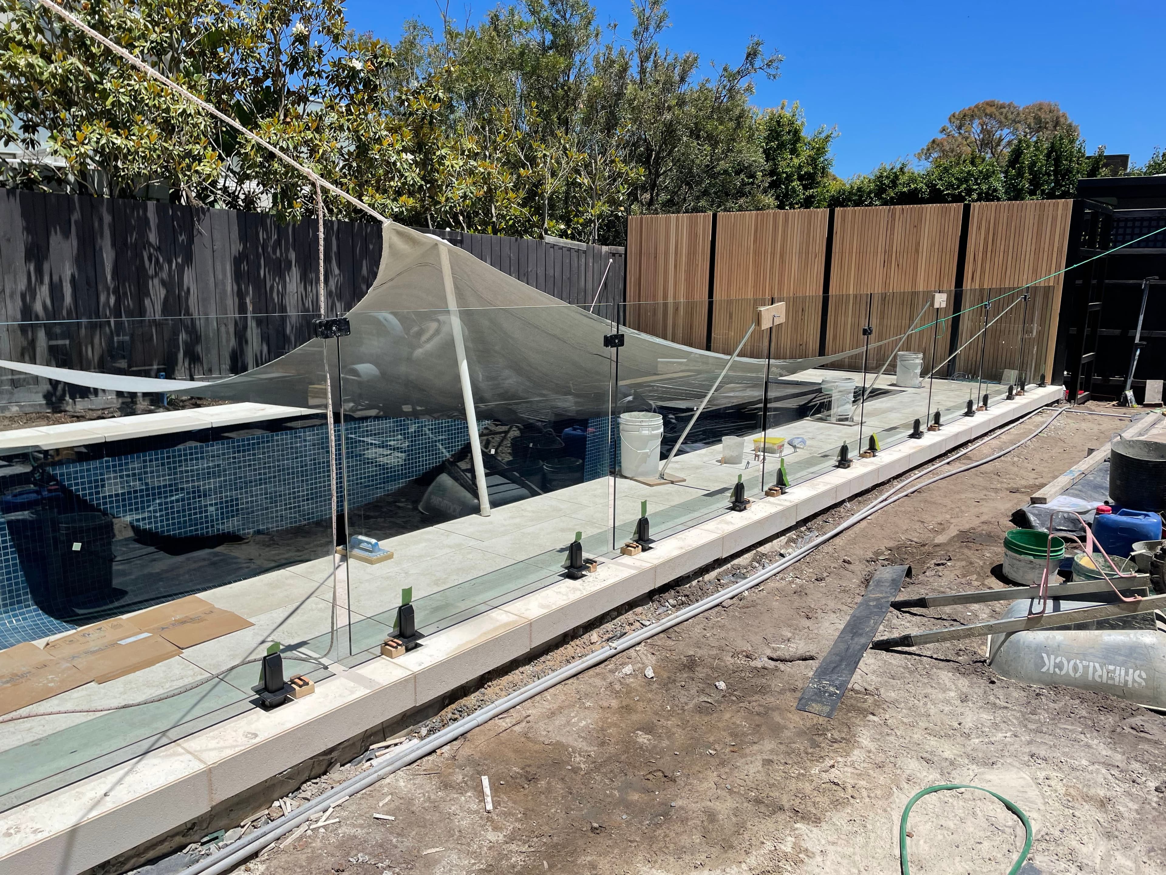 Frameless glass pool fence with matte black spigots core-drilled into pool coping surrounding a tiled swimming pool under construction in a suburban backyard.