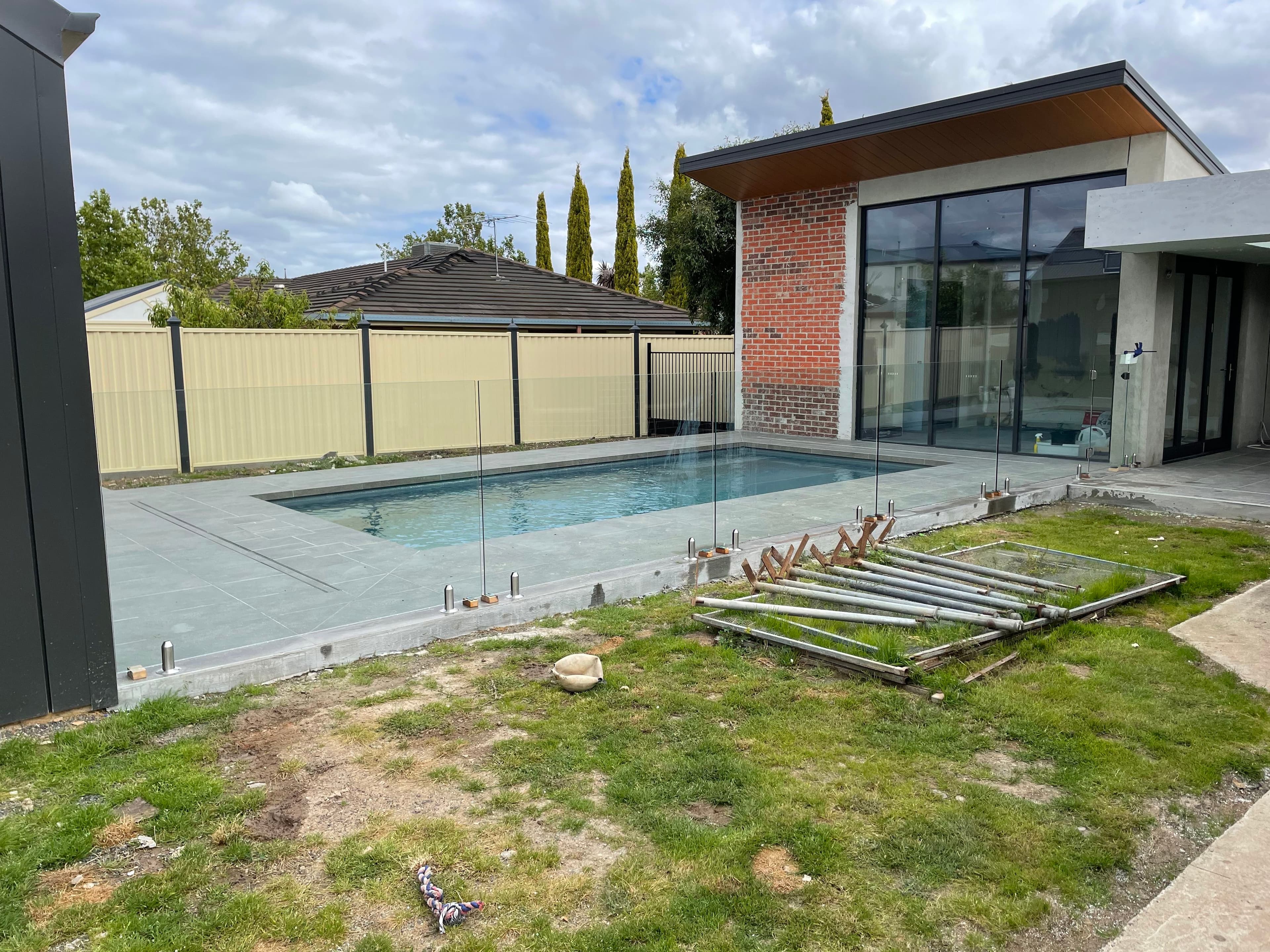 Frameless glass pool fence with chrome spigots core-drilled into concrete coping surrounding a backyard swimming pool, with glass panels partially installed during construction at a modern Victorian home.