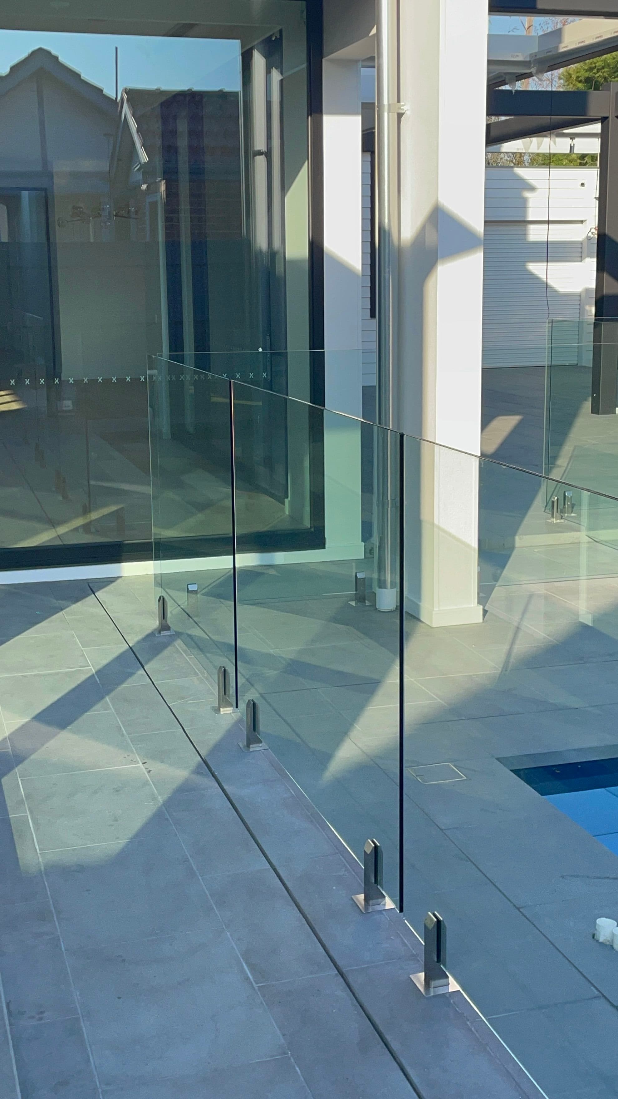 Frameless pool fence with brushed nickel square spigots surface-mounted on grey stone coping, clear glass panels surrounding a backyard pool with visible blue water at lower right.