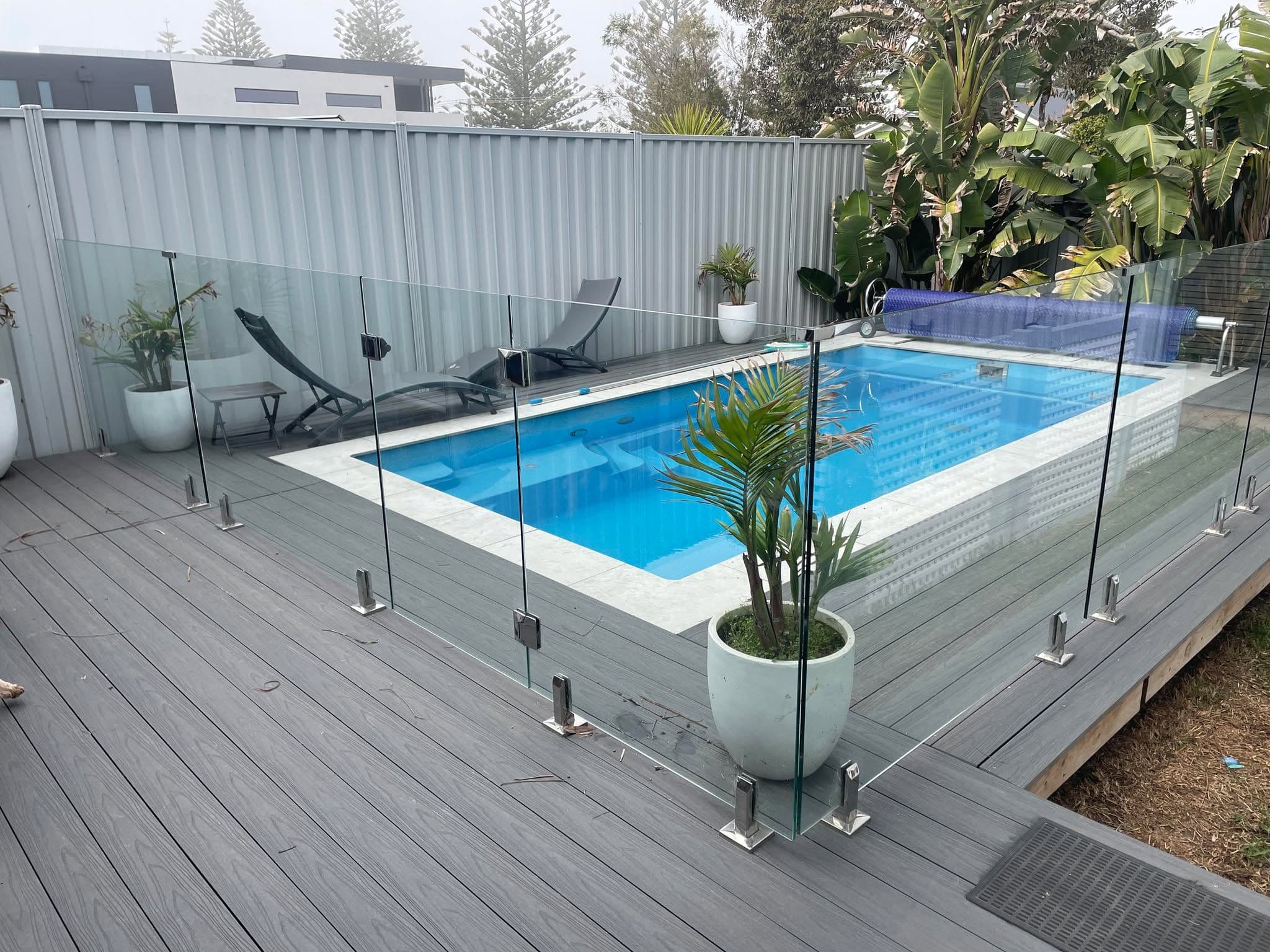 Frameless glass pool fence with chrome spigots surface-mounted into grey composite decking surrounding a backyard swimming pool with white coping.