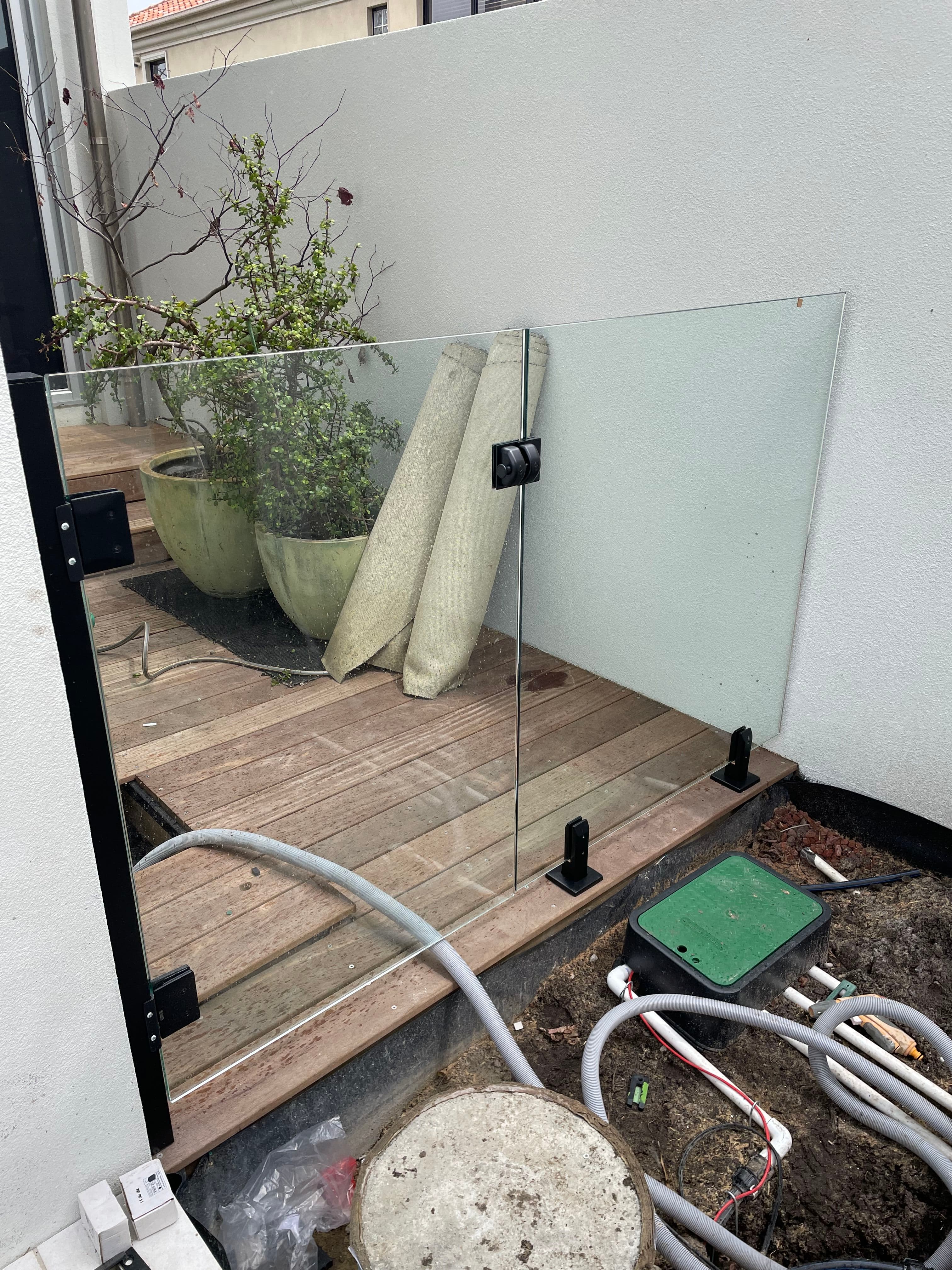 Frameless glass pool fence panel with matte black square spigots fixed to timber decking during installation, with no pool water visible in the image.