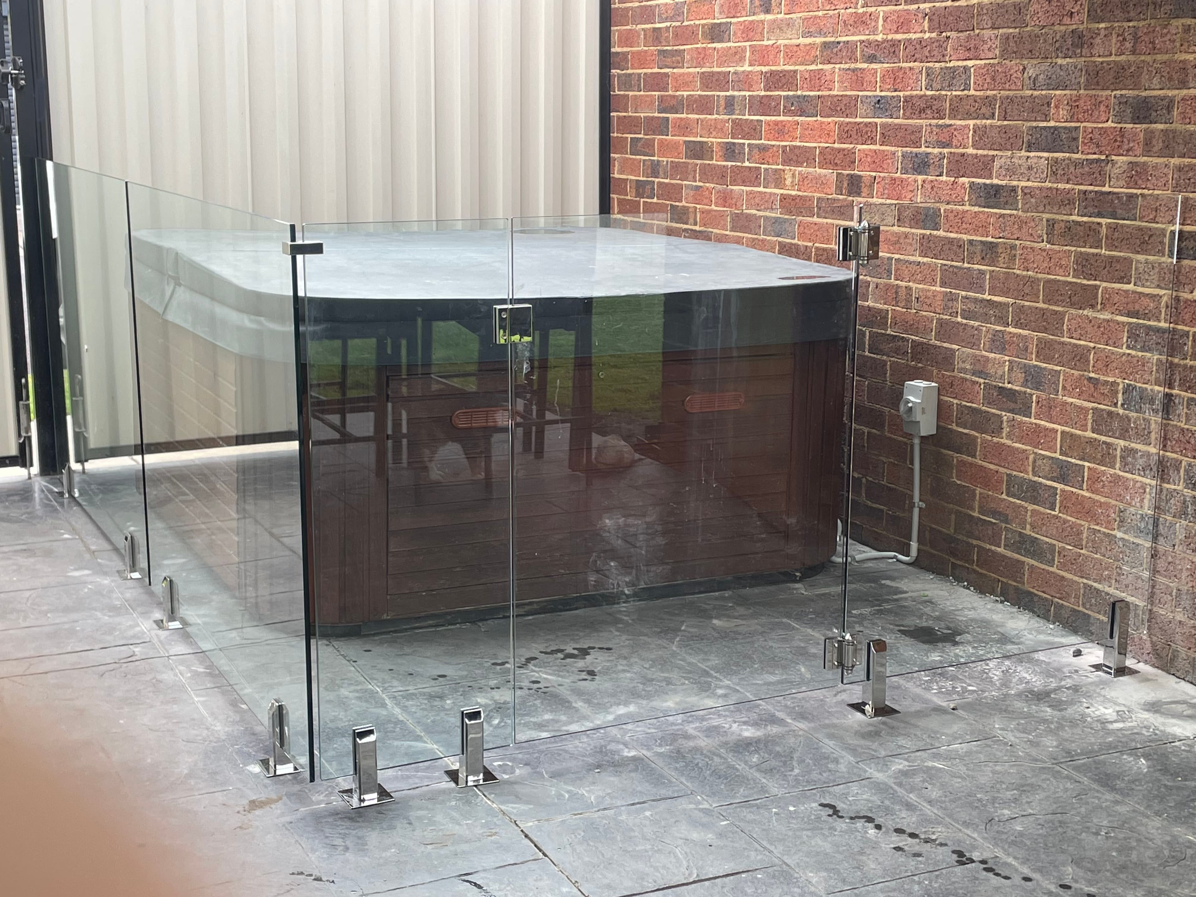 Frameless pool fence with chrome spigots surface-mounted to tiled paving, enclosing what appears to be a spa or pool structure with a marble-look coping edge, set against a brick wall exterior.