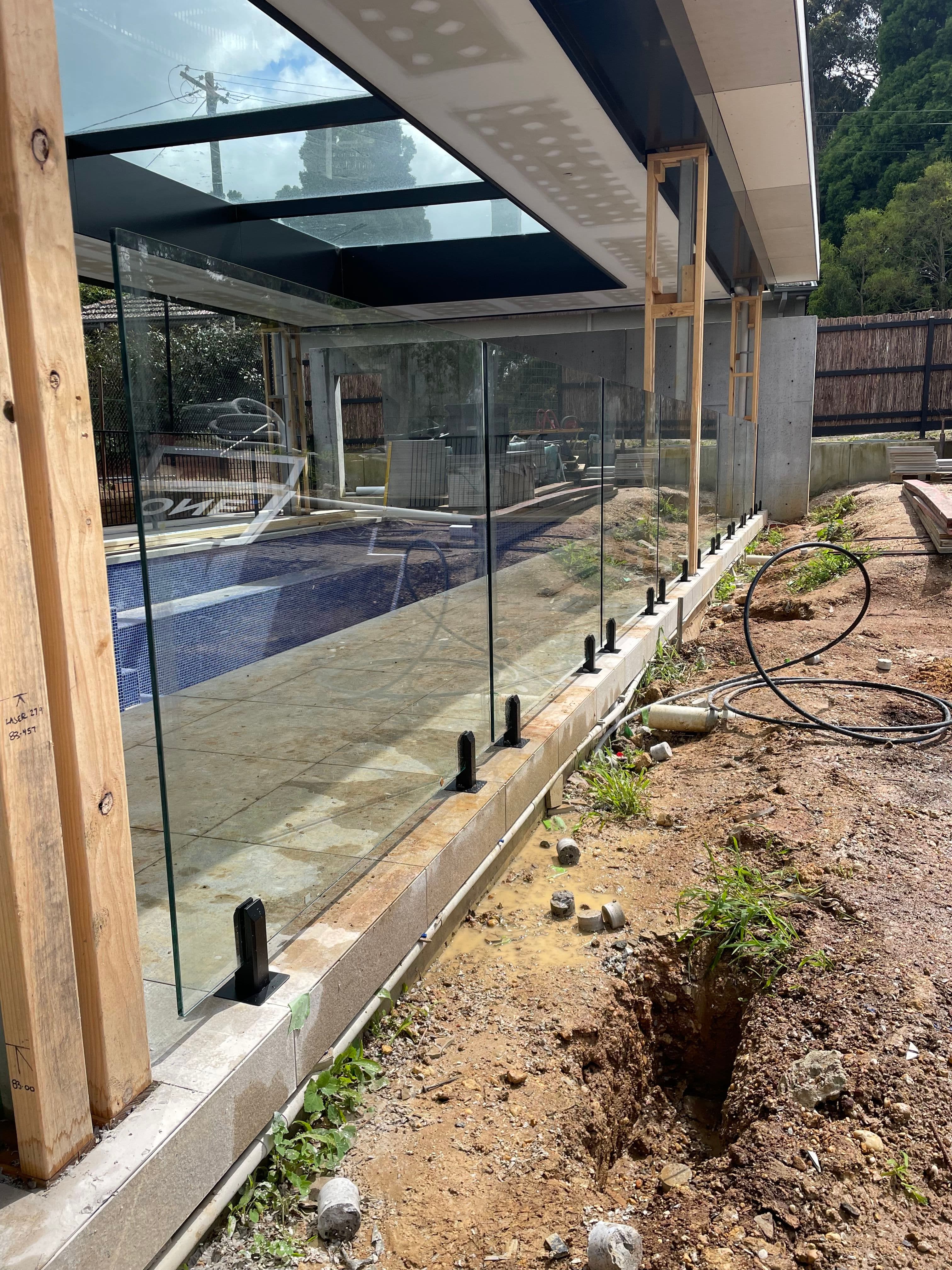 Frameless pool fence with matte black spigots core-drilled into a concrete coping wall surrounding an under-construction backyard swimming pool with visible blue tiled pool water.