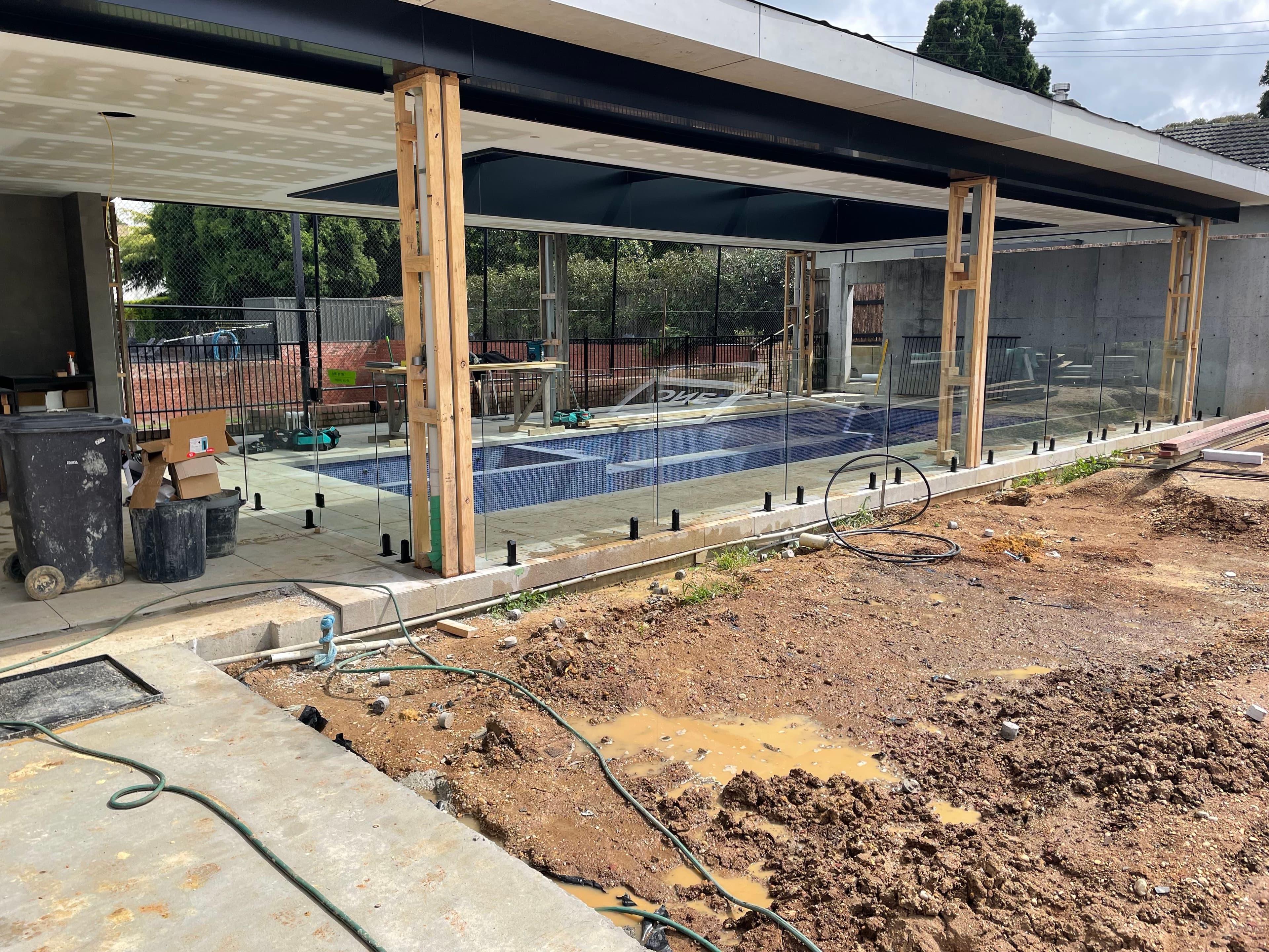 Frameless glass pool fence with matte black spigots installed around a tiled swimming pool during construction, set in a residential outdoor alfresco area under a modern pergola structure.