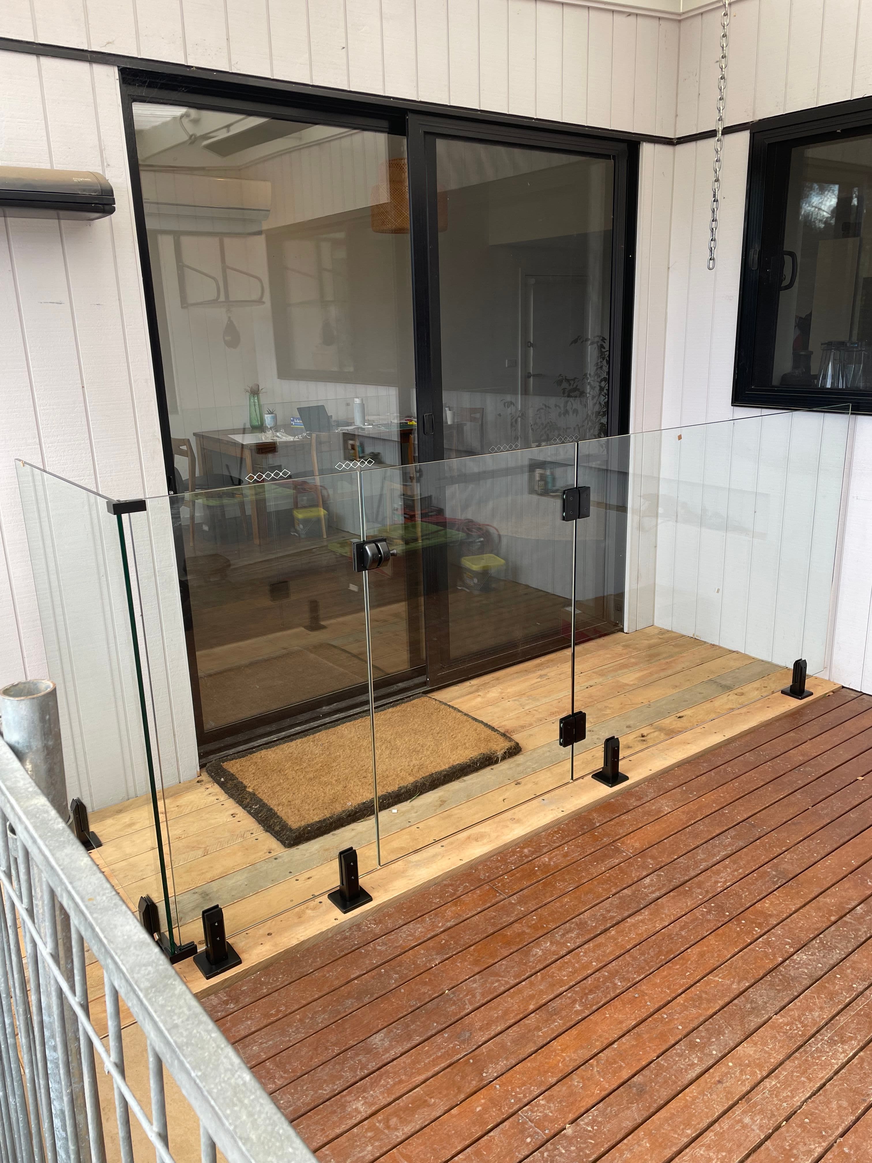 Frameless glass balustrade panels with matte black square spigots surface-mounted on a timber deck in front of a sliding door entry, with no pool visible.