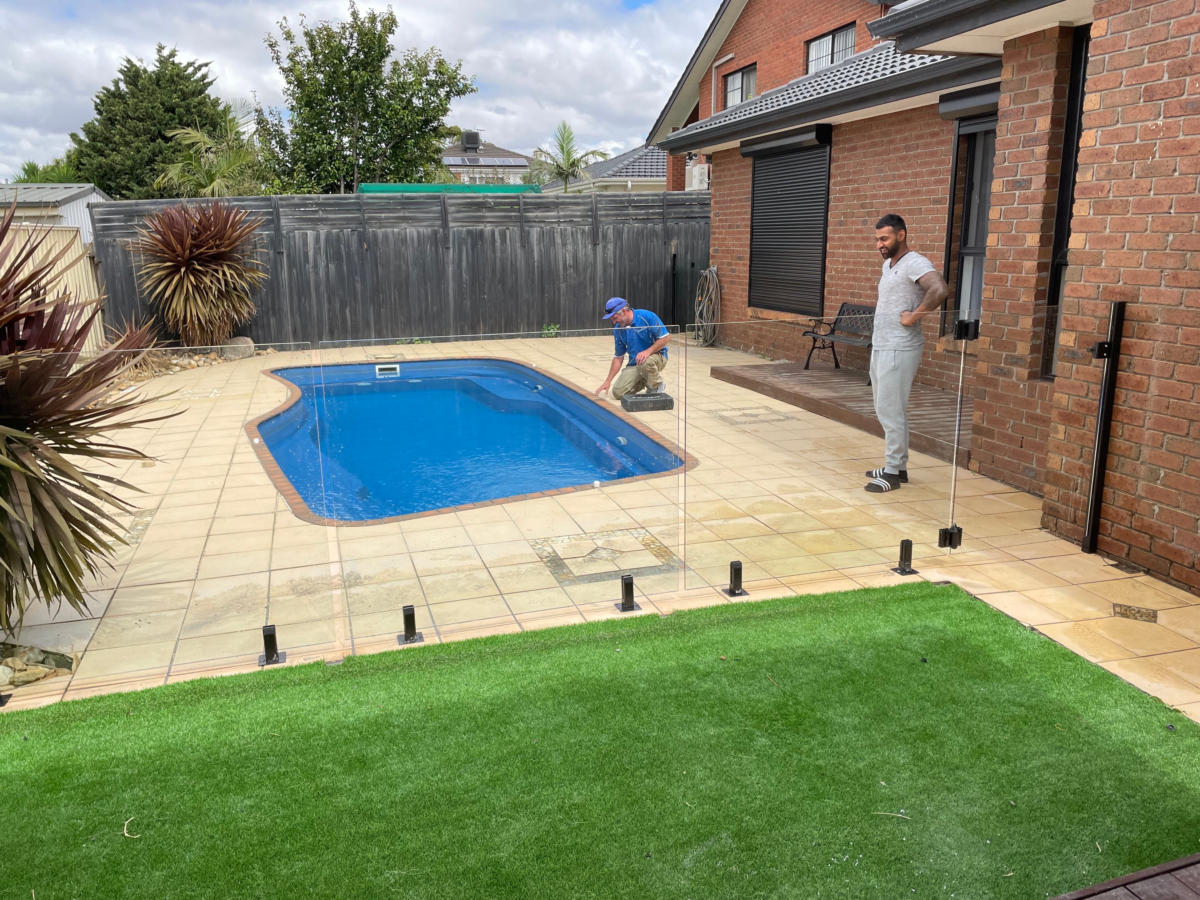 Frameless glass pool fence with matte black spigots core-drilled into paved coping surrounding a backyard swimming pool, with two installers fitting the panels during installation.