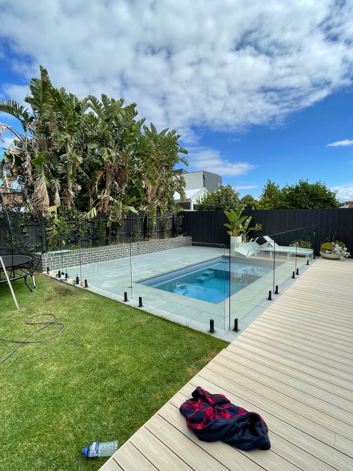 Frameless pool fence with matte black spigots core-drilled into stone coping surrounding a rectangular backyard pool, with timber decking and lush tropical landscaping.