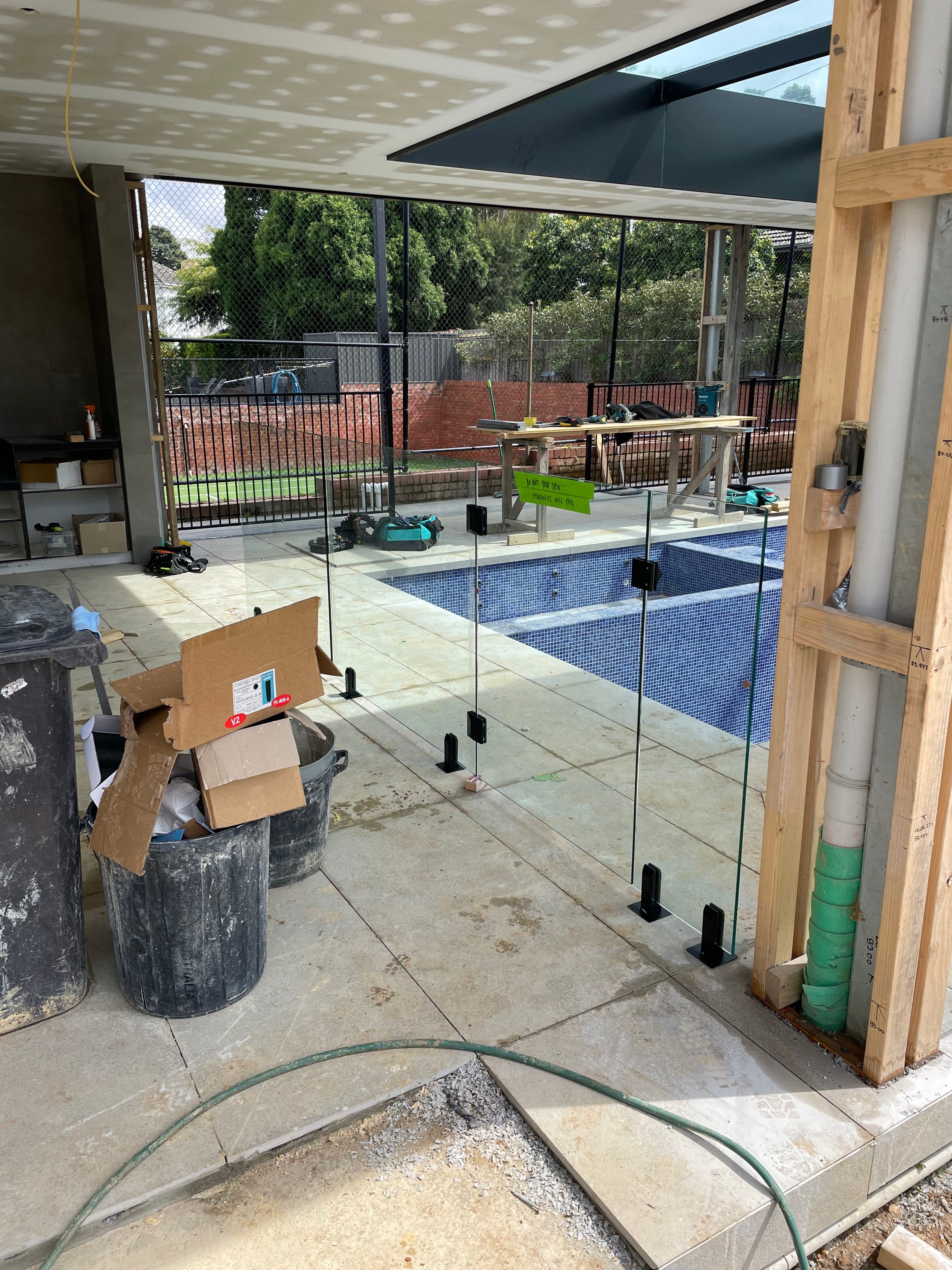 Frameless glass pool fence with matte black spigots core-drilled into concrete coping surrounding an in-construction backyard swimming pool with blue mosaic tiles visible.