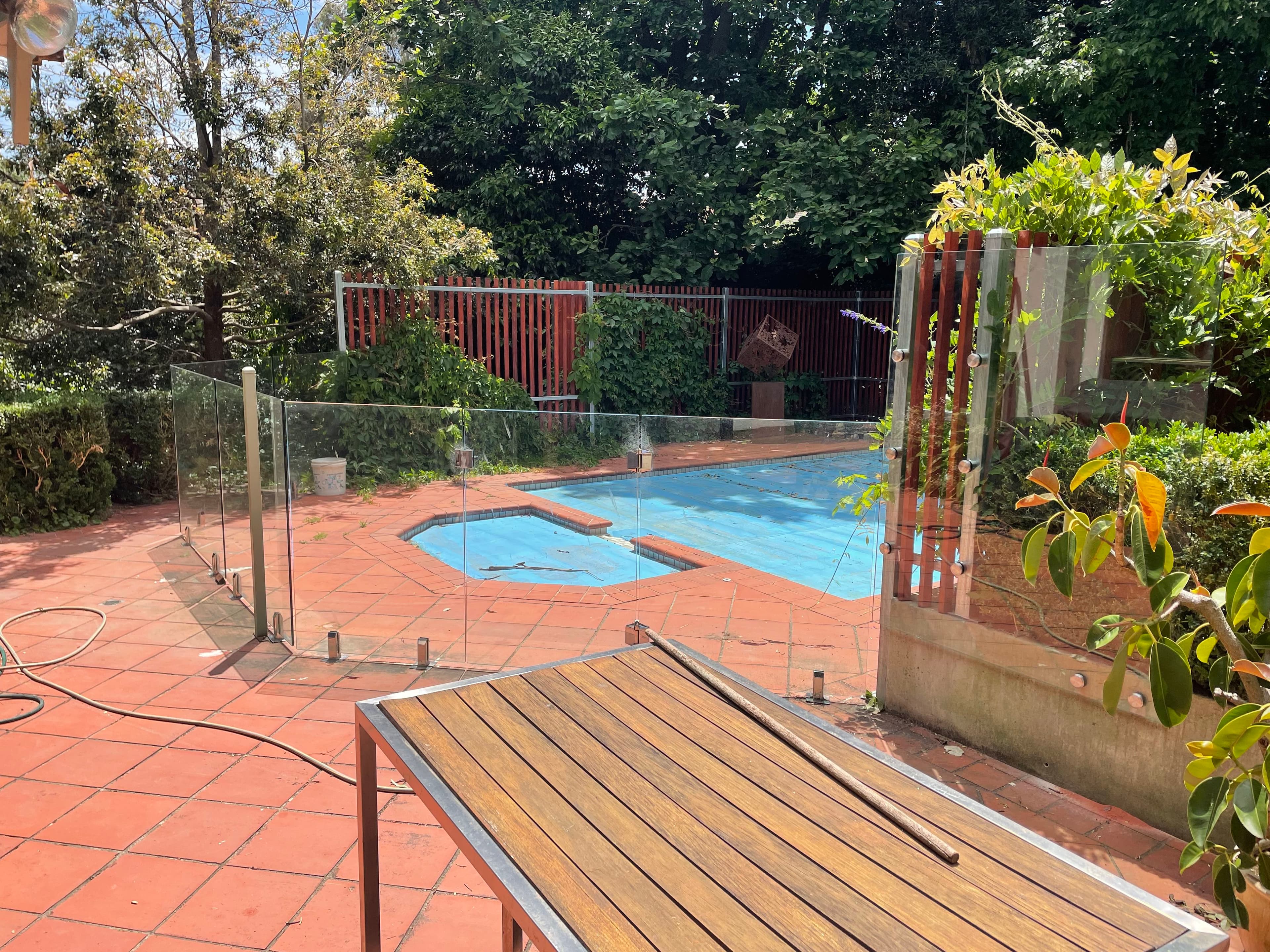 Frameless glass pool fence with chrome spigots core-drilled into terracotta paving surrounding a backyard swimming pool with visible blue water.