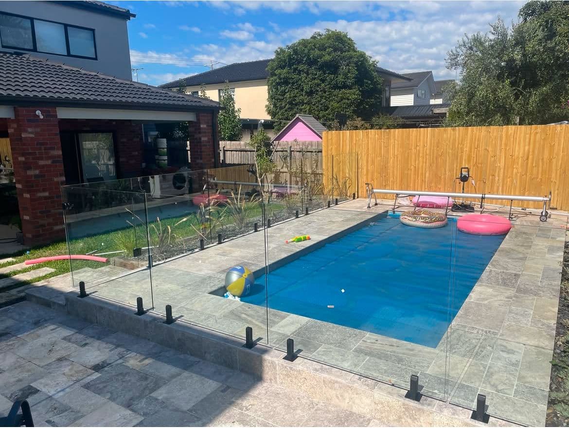 Frameless glass pool fence with matte black square spigots core-drilled into stone coping surrounding a rectangular backyard swimming pool in a suburban Melbourne home.