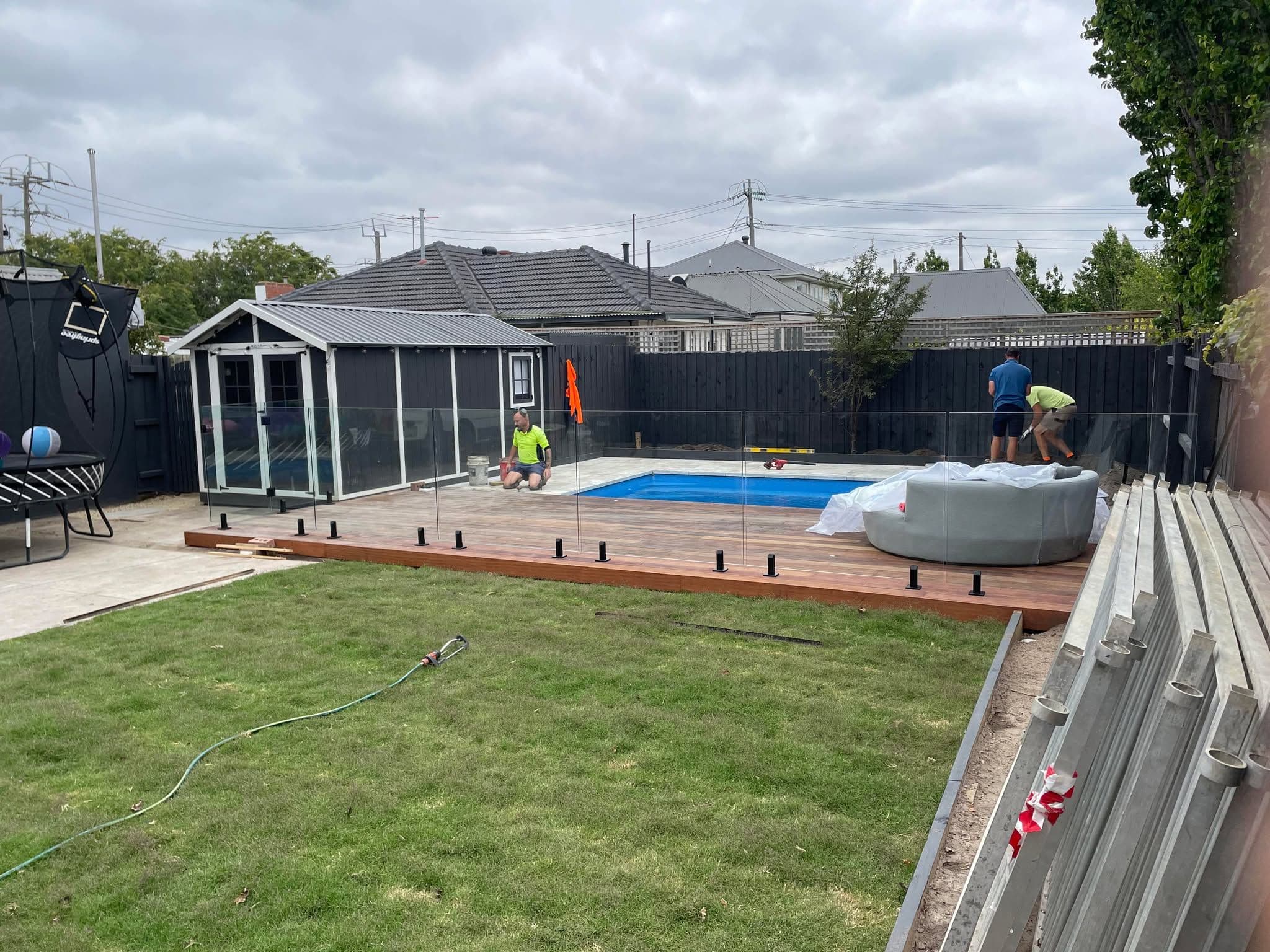 Frameless glass pool fence with matte black spigots being installed around a backyard swimming pool on a timber deck, with pool water clearly visible.