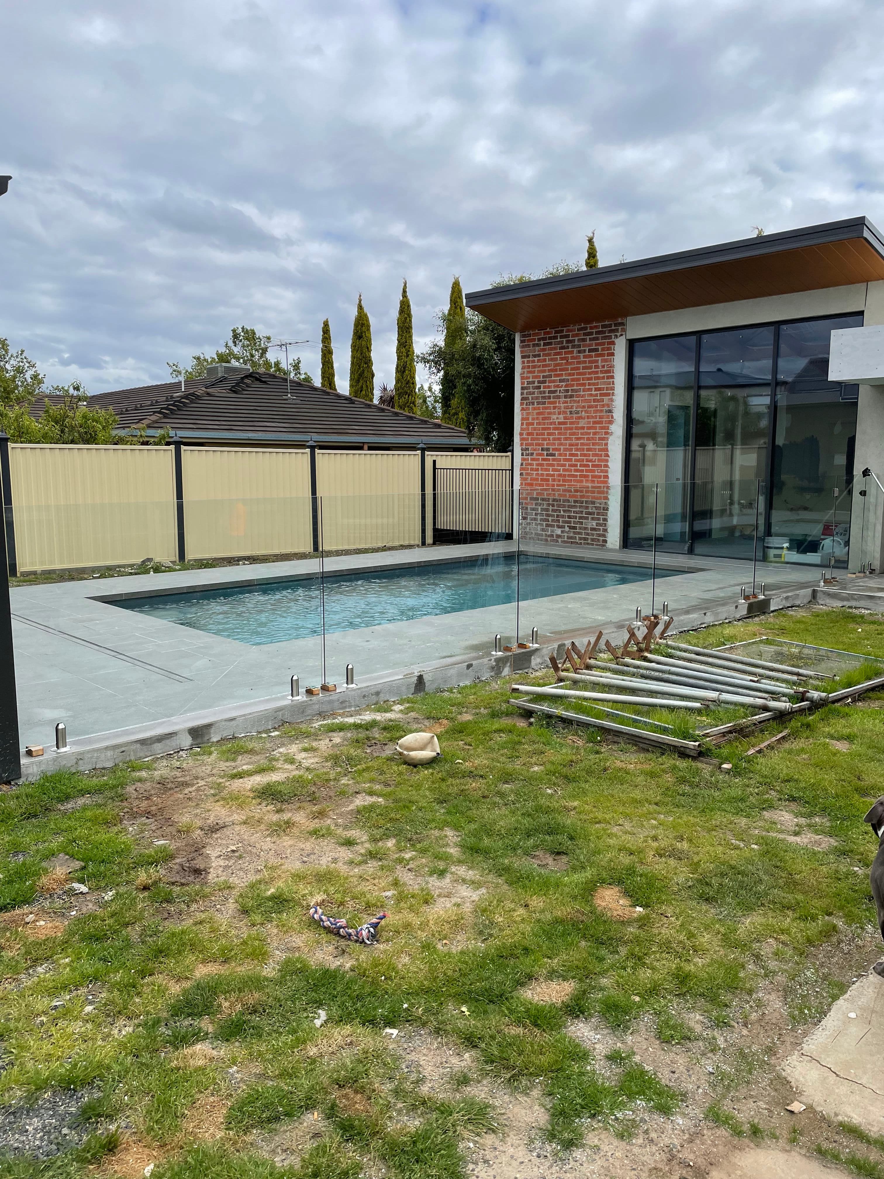 Frameless glass pool fence with chrome spigots core-drilled into concrete coping surrounding a rectangular backyard swimming pool beside a modern brick and timber home.