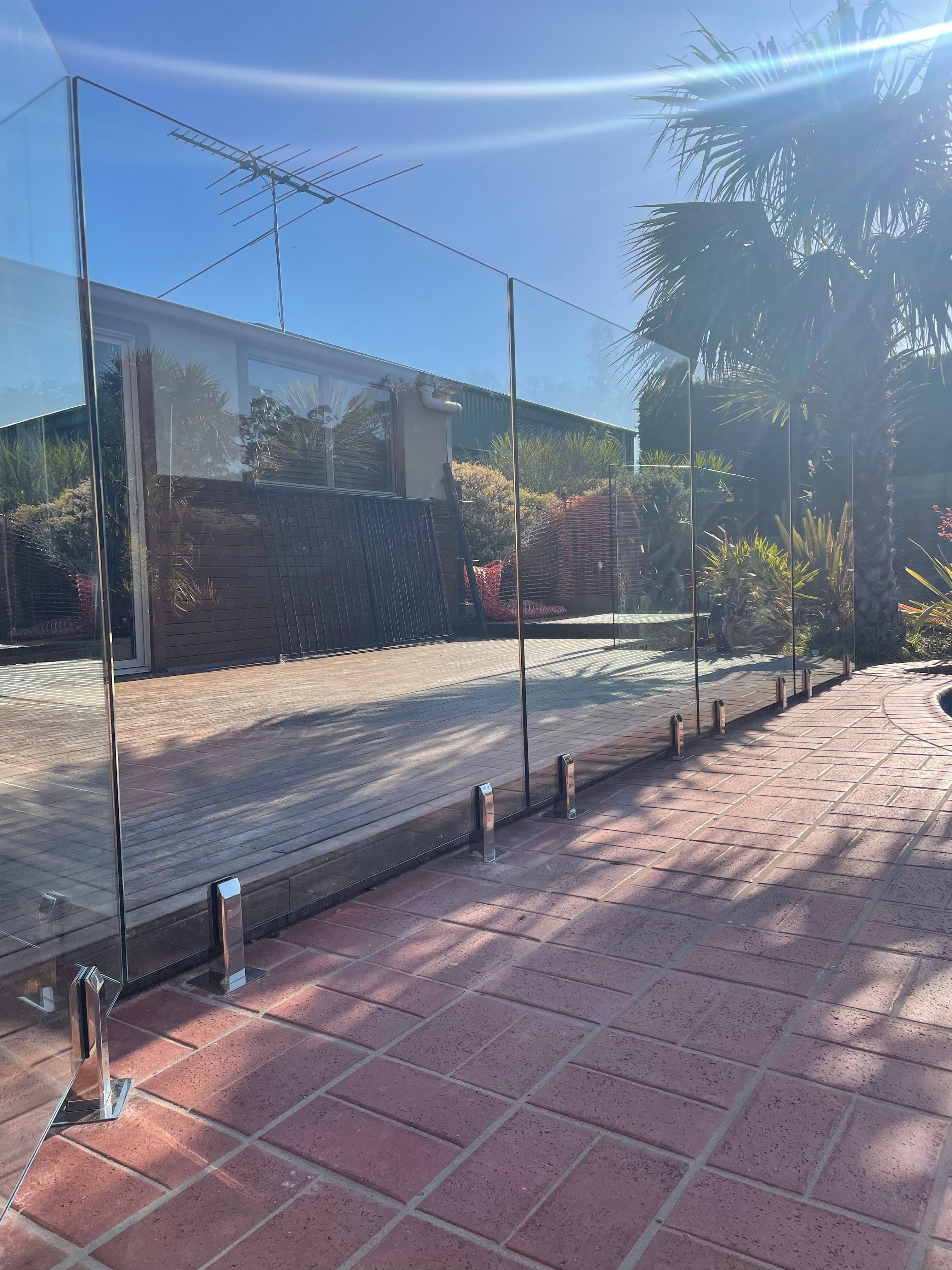 Frameless glass pool fence with chrome spigots core-drilled into brick paving and timber decking edging, surrounding an outdoor alfresco area with tropical landscaping.