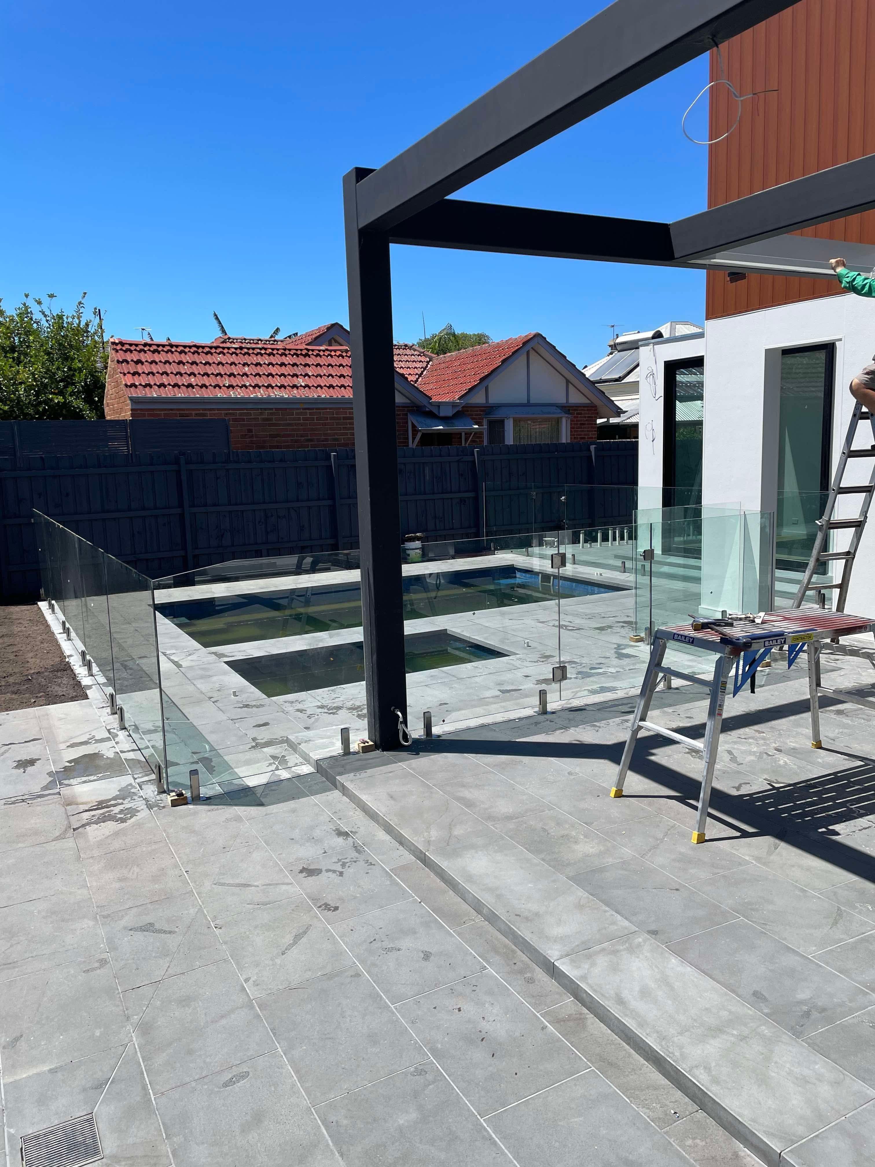 Frameless pool fence with chrome spigots core-drilled into stone coping surrounding a backyard swimming pool, during installation on a modern home with a black steel pergola.