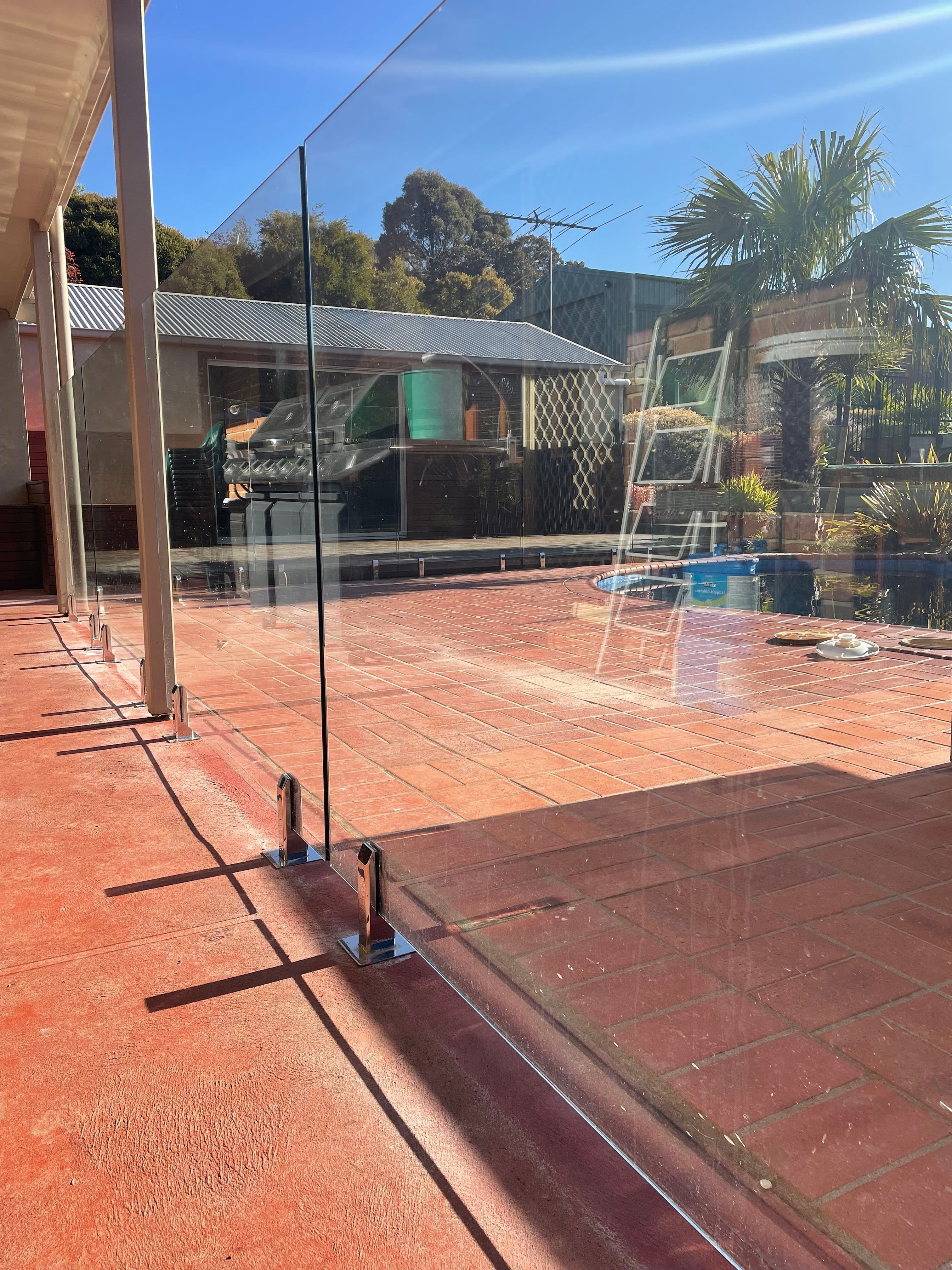 Frameless pool fence with chrome spigots core-drilled into red brick paving surrounding a backyard pool, with clear glass panels and a suburban outdoor setting visible.