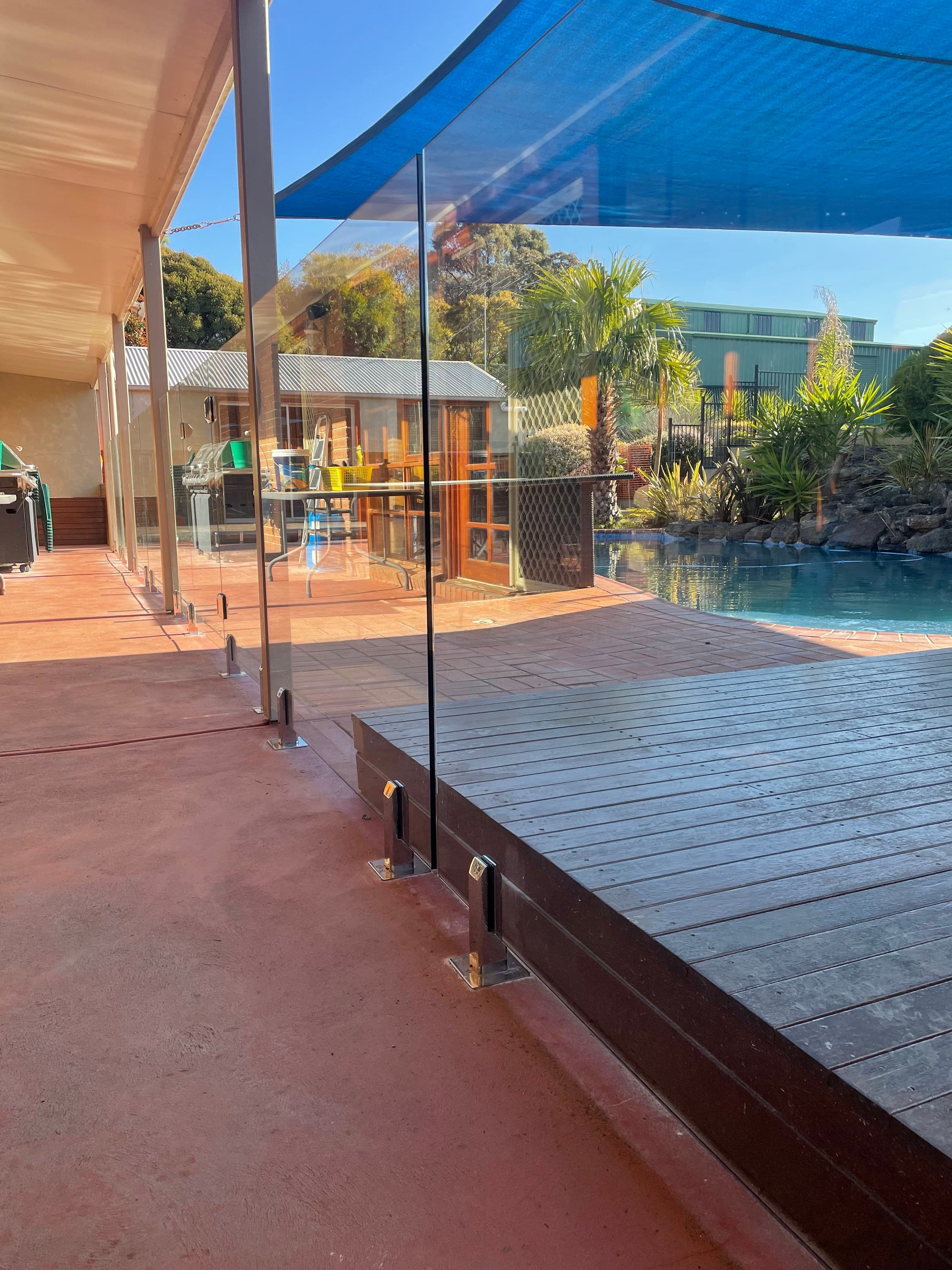 Frameless pool fence with chrome square spigots core-drilled into concrete and timber decking surrounds a backyard swimming pool with visible blue water, adjacent to an outdoor entertaining area.
