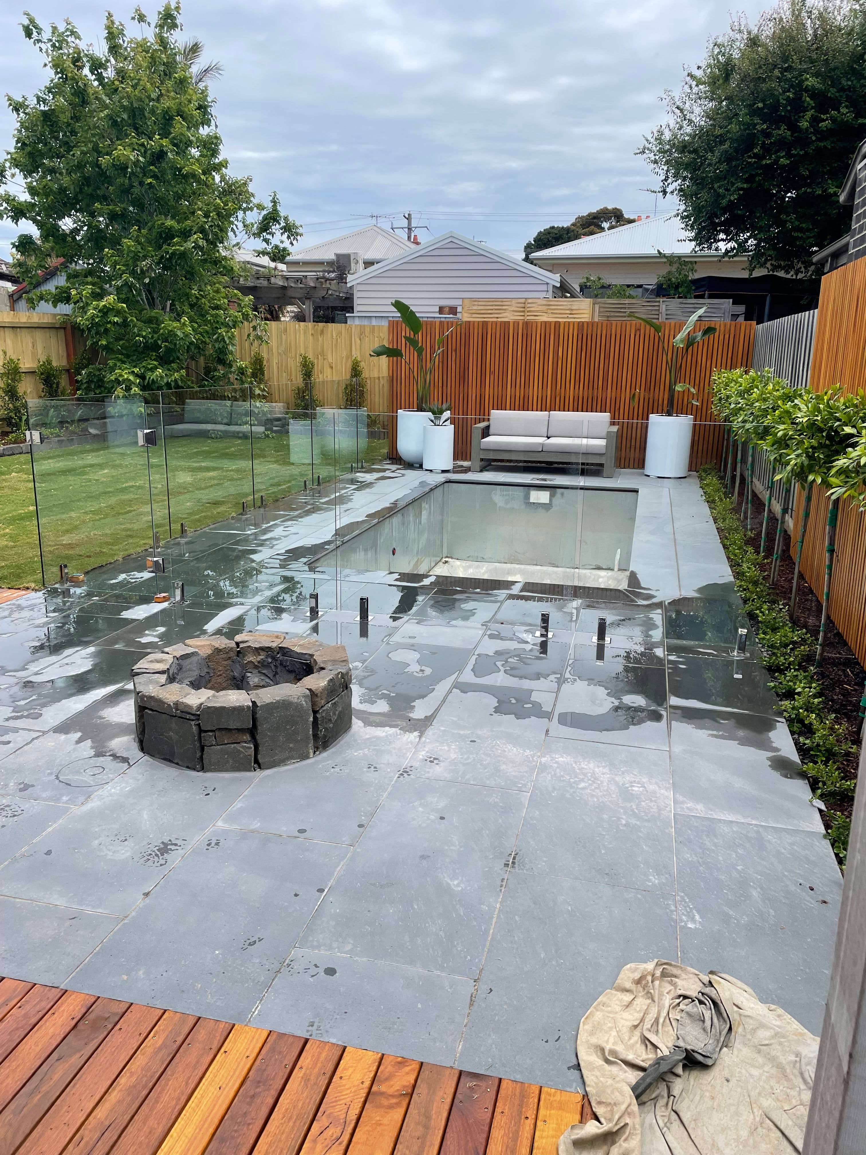 Frameless glass pool fence with matte black spigots core-drilled into bluestone coping surrounding a rectangular backyard swimming pool, with timber decking and a stone fire pit visible in the foreground.