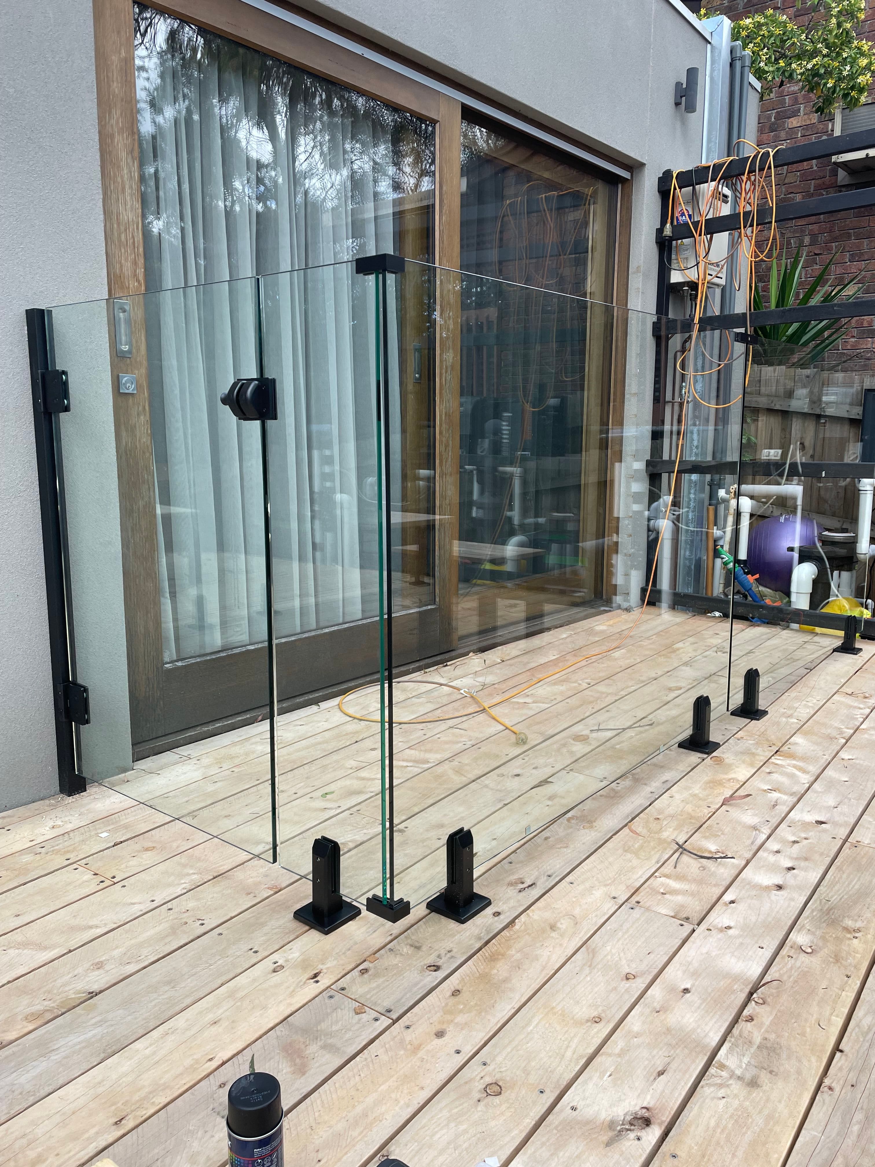 Frameless glass balustrade panels with matte black square spigots installed on a timber deck adjacent to a house with sliding doors — no pool water or pool coping visible.