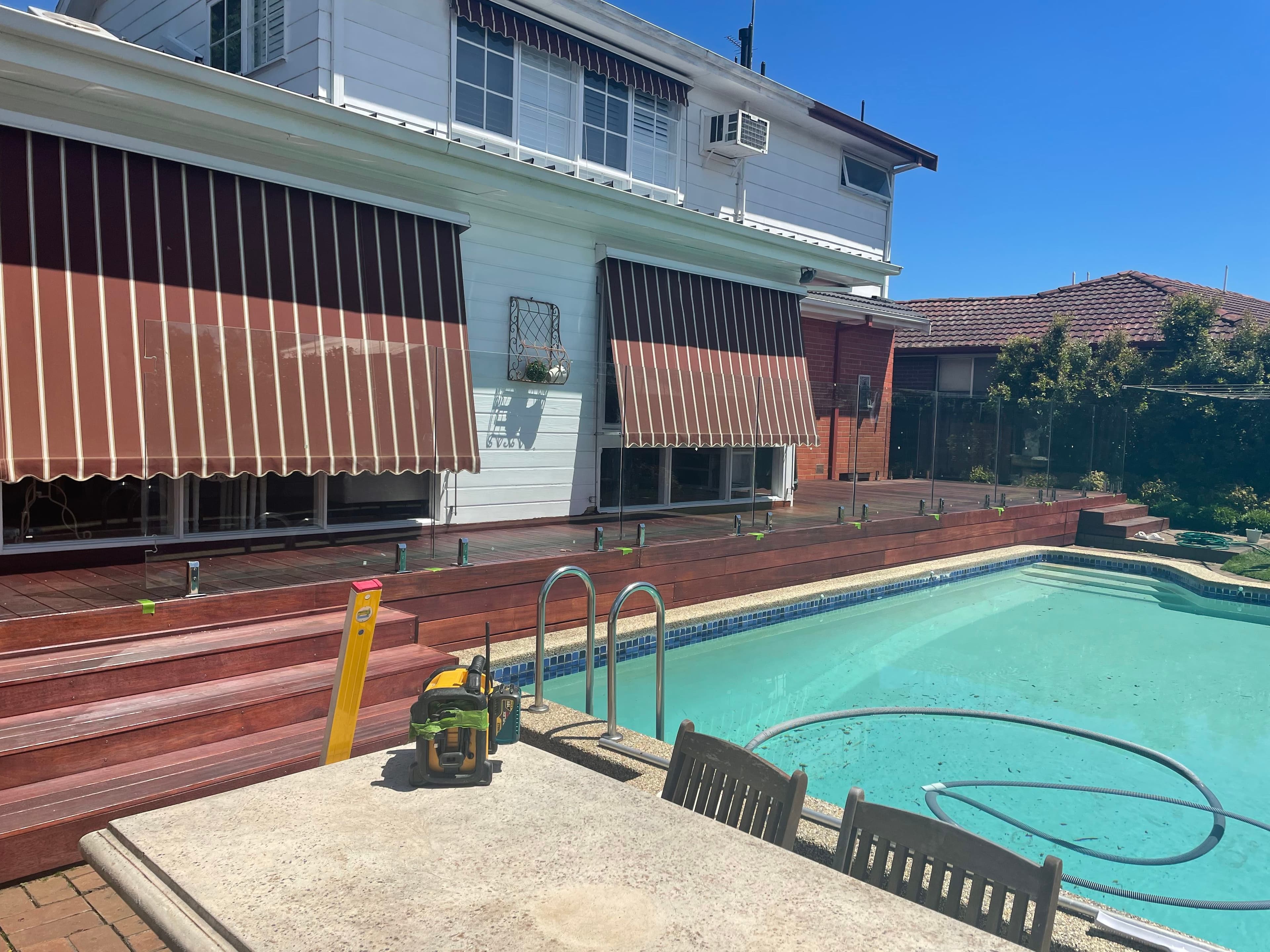 Frameless glass pool fence being installed with spigots core-drilled into a timber deck surrounding a backyard swimming pool, adjacent to a white weatherboard home with striped awnings.