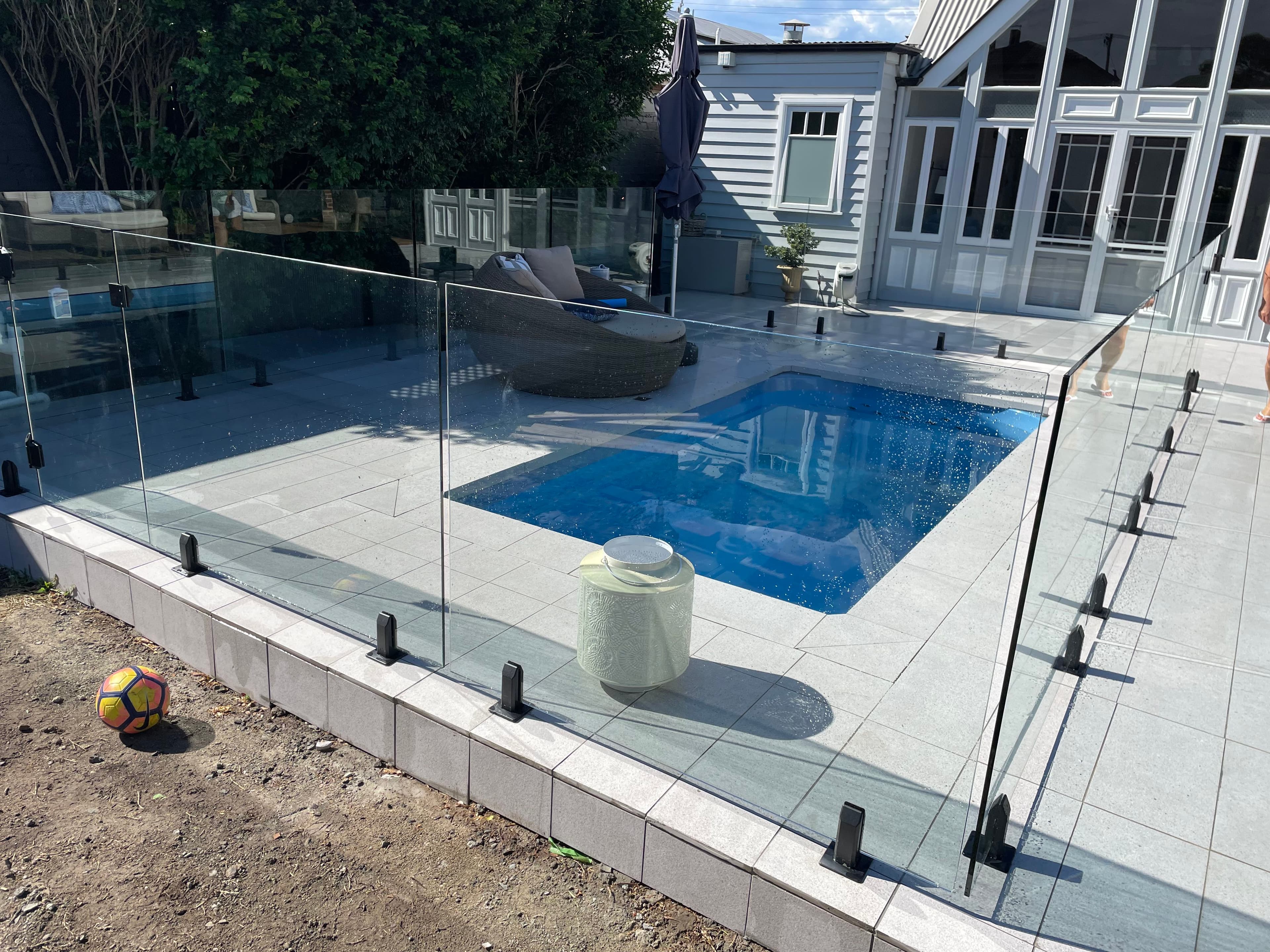 Frameless pool fence with matte black square spigots surface-mounted into grey stone coping surrounding a small rectangular backyard pool adjacent to a classic weatherboard home.