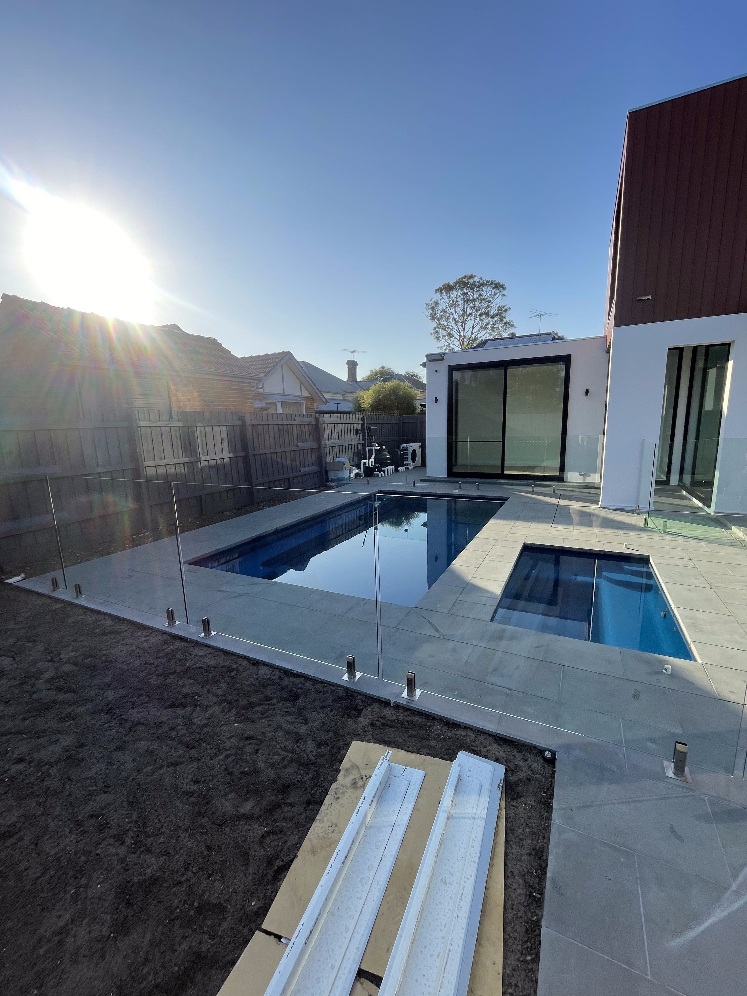 Frameless glass pool fence with chrome spigots core-drilled into concrete coping surrounding a rectangular backyard swimming pool and spa, adjacent to a modern contemporary home.