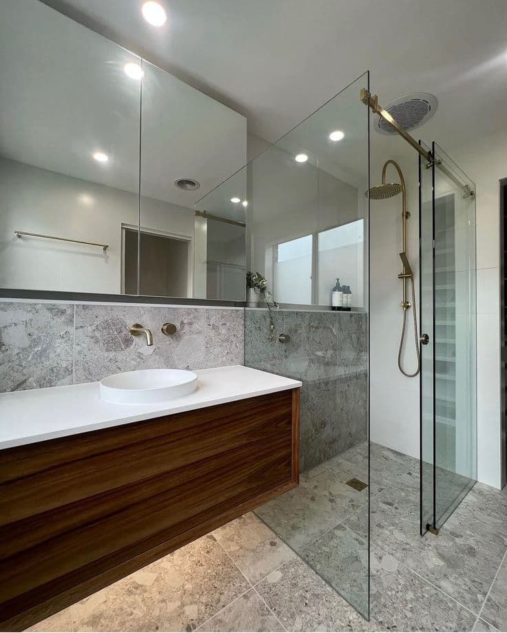 Semi-frameless shower enclosure with a brushed gold top rail and sliding door track in a marble-tiled bathroom with matching brushed gold tapware and a floating timber vanity.