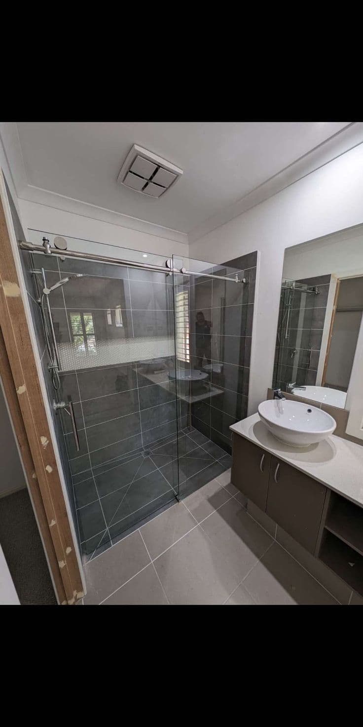 Semi-frameless sliding shower screen with a visible satin chrome top rail and sliding track in a dark grey tiled bathroom with a vessel sink vanity.
