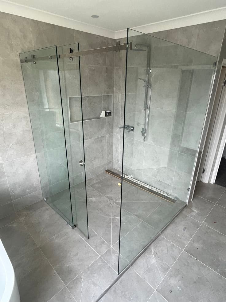 Semi-frameless shower enclosure with a visible chrome sliding door top rail and track hardware in a corner configuration, set in a grey marble-look tiled bathroom.