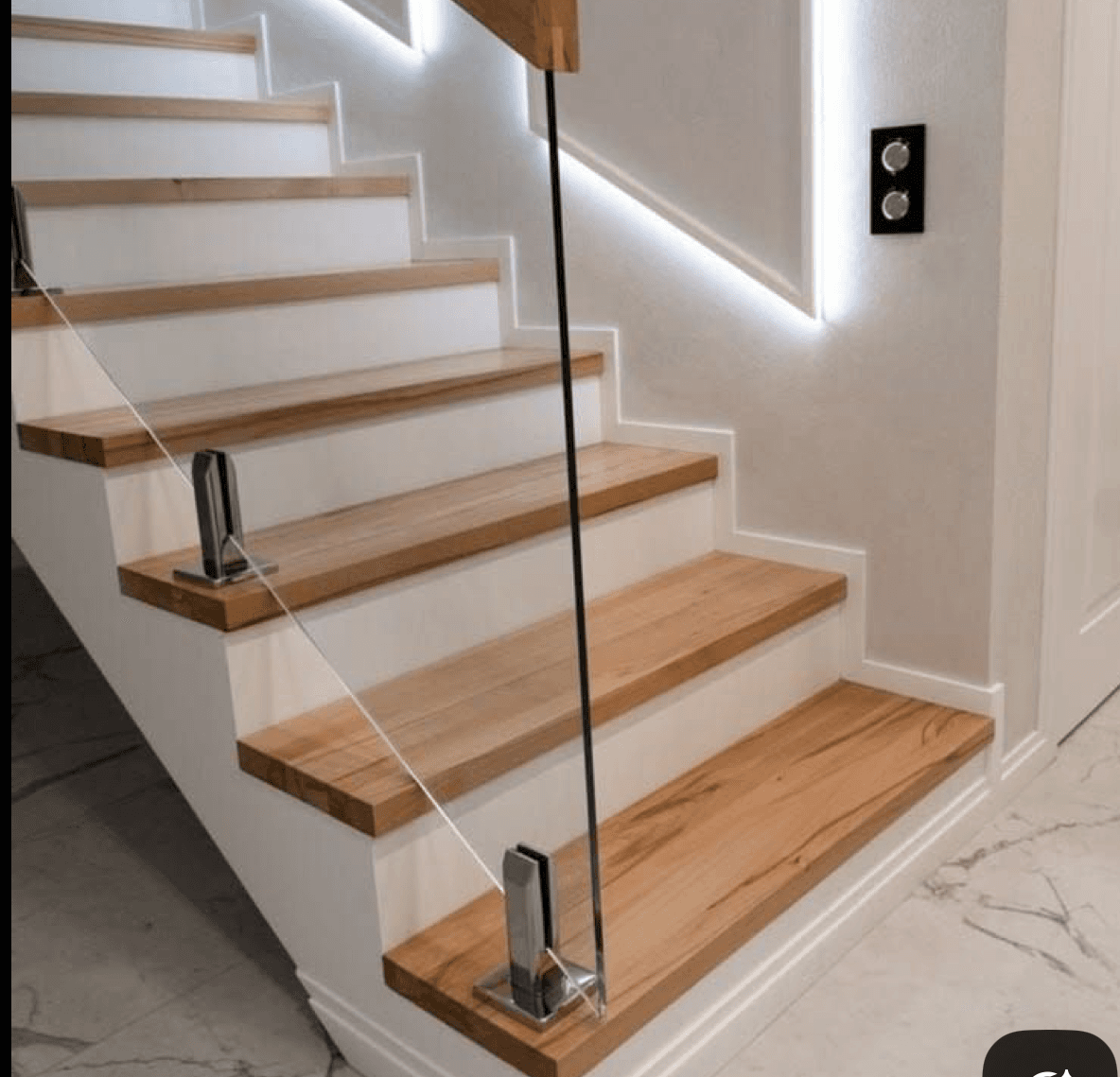 Clear glass stair balustrade with chrome spigots fixed to timber stair treads in a modern interior with LED strip lighting and marble flooring.